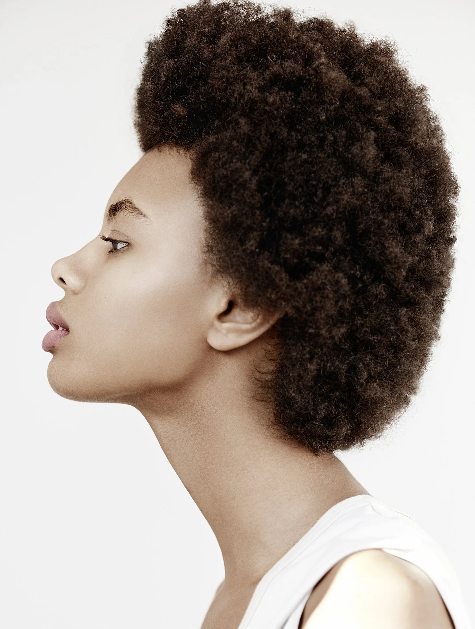 Seitenansicht eines jungen Mannes mit lockigem, dunklem Haar und glatter Haut, in einem weißen Oberteil, vor weißem Hintergrund.