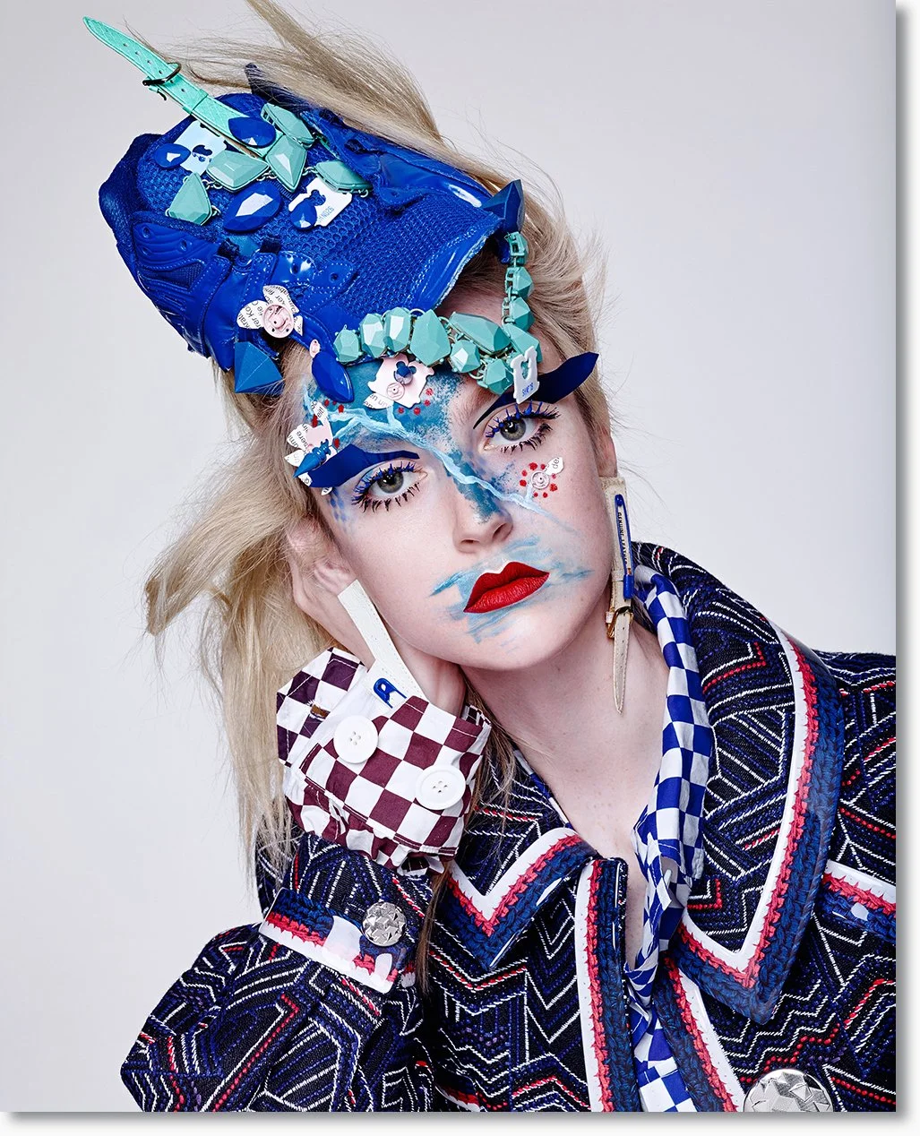 Porträt einer Frau mit auffälligem, kreativen Make-up und Accessoires: Sie trägt einen bunten, gemusterten Anzug, große Ohrringe und ein auffälliges Kopfstück aus blauen und türkisfarbenen Objekten, die an Geode und Kristalle erinnern. Ihr Make-up is