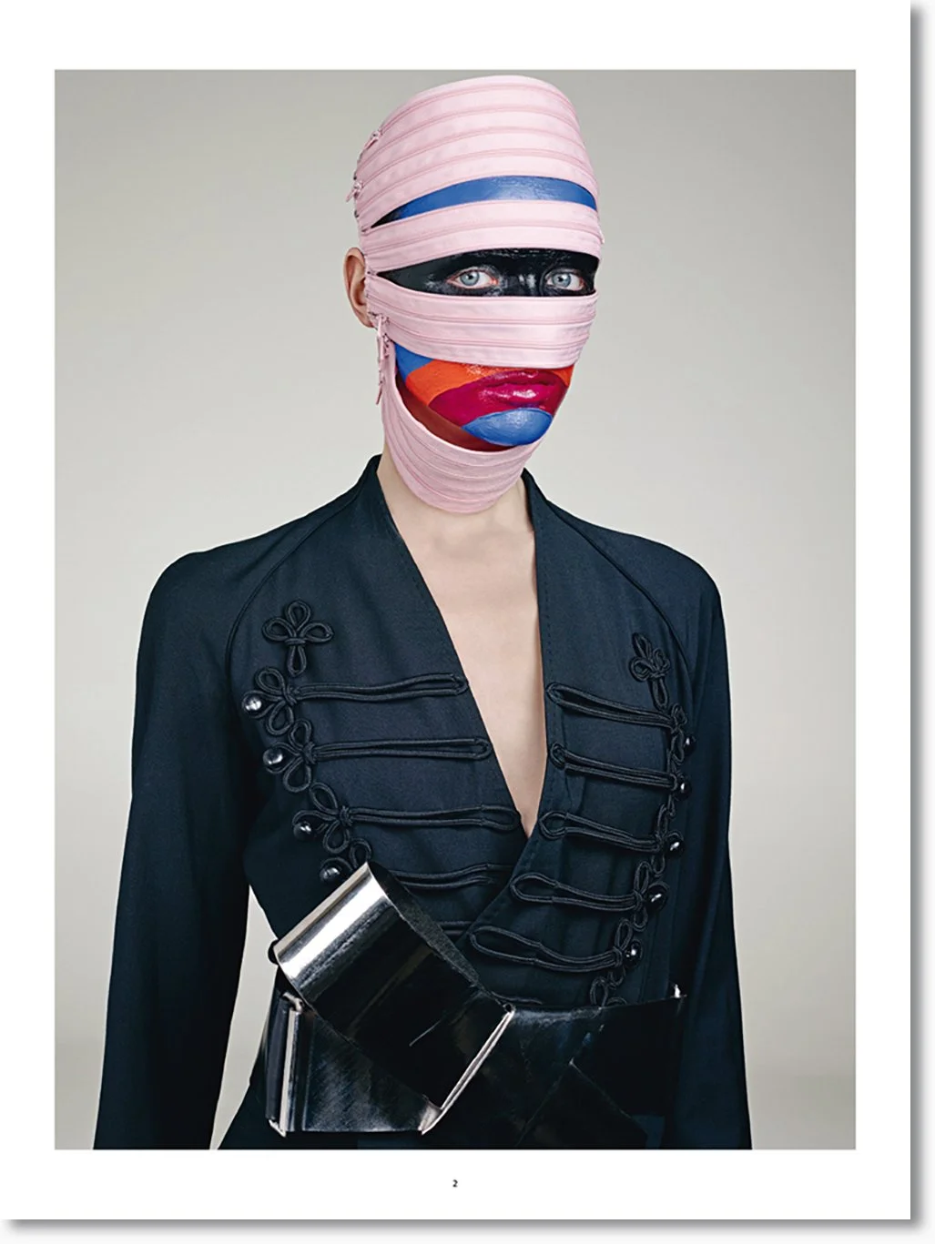 Person trägt ein kreatives Gesichtsmasken-Design aus Bändern mit buntem Lippen- und Augendruck, schwarze Jacke mit dekorativen Knoten, und metallic-artiges Accessoire. Hintergrund ist schlicht.
