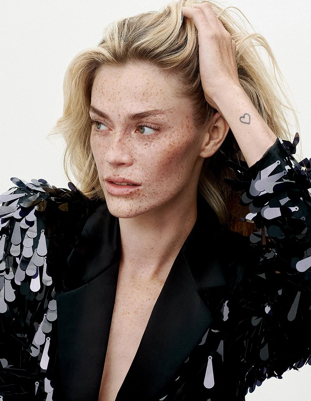 Nahaufnahme einer Frau mit blonden, lockigen Haaren und Sommersprossen. Sie trägt ein schwarzes Outfit mit einer Schulterpartie, verziert mit schwarzen Pailletten, und hat ein kleines Herz-Tattoo am Handgelenk.