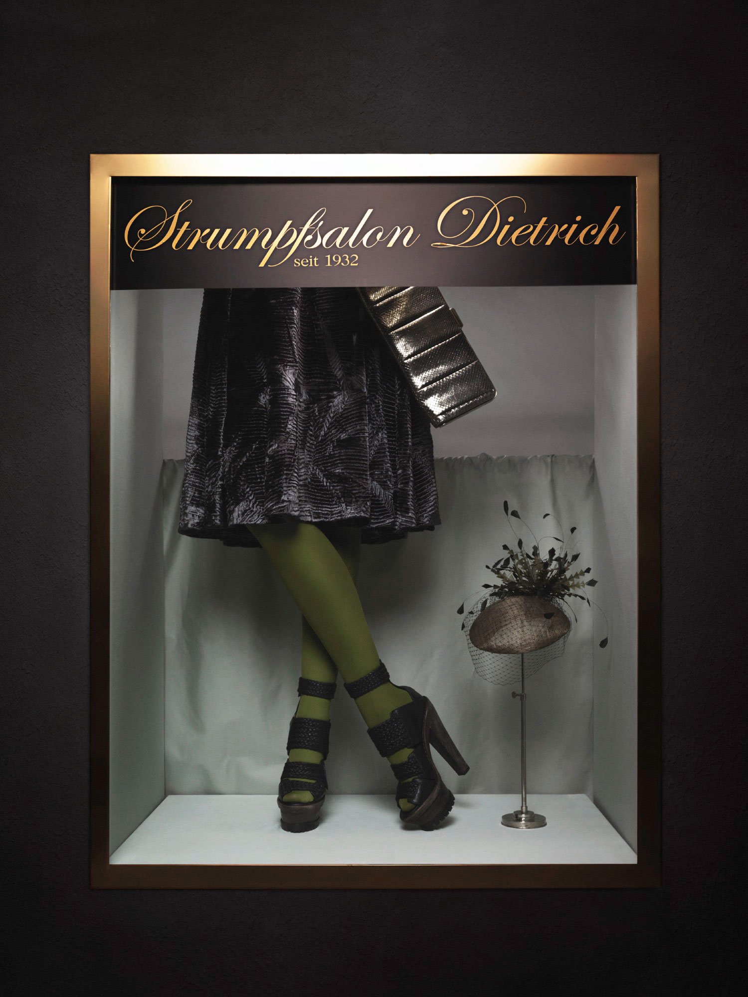 Display of a mannequin's legs in green tights, wearing black high-heeled shoes with straps, in a glass case at a fashion exhibit for Strumpfsalon Dietrich. The upper part shows a black skirt and a black metallic bag, with a sign that reads "Strumpfsa