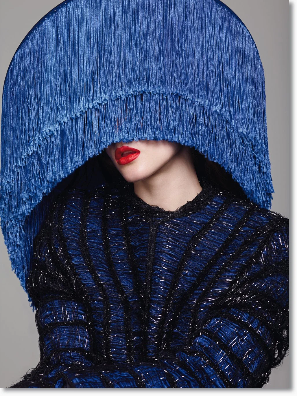 Frau trägt einen avantgardistischen blauen Hut mit Fransen, der ihre Augen verdeckt, und ein passendes Outfit aus schwarzem, wabenförmig strukturiertem Material mit blauen Akzenten. Sie trägt rote Lippen.