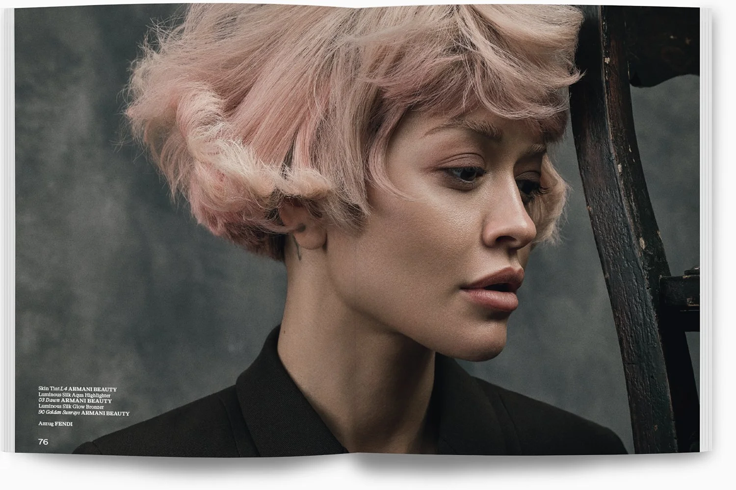 Nahaufnahme eines Models mit kurzen, gewellten, rosafarbenen Haaren, das nach unten schaut. Der Hintergrund ist grau, und das Model trägt einen schwarzen Blazer.