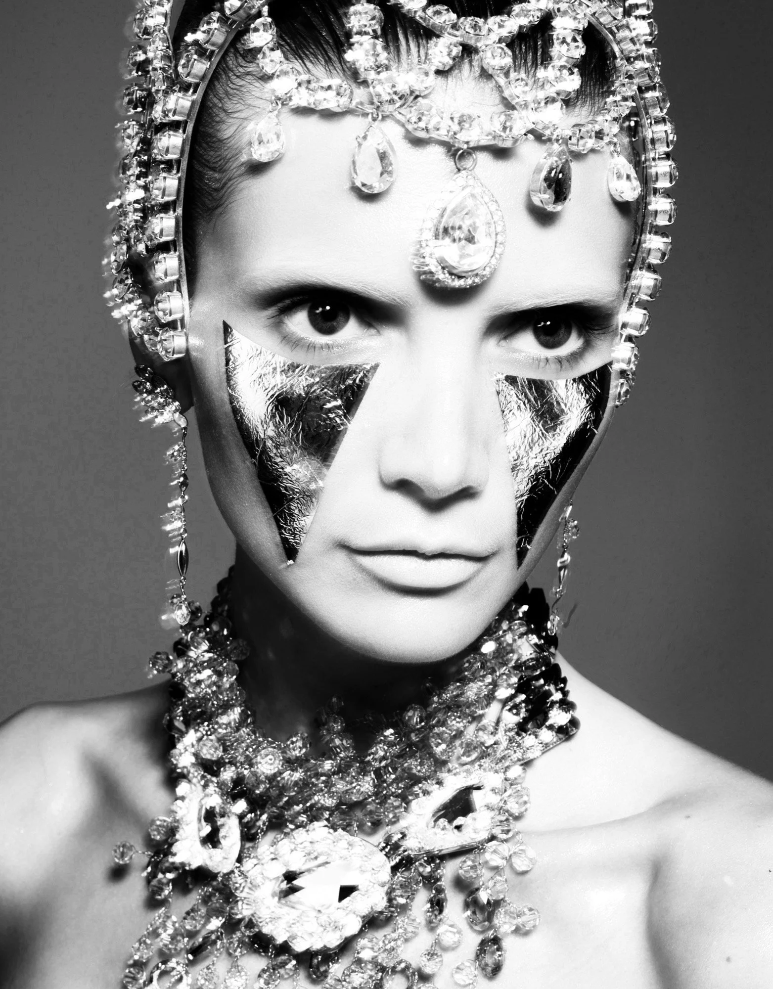 A woman wearing an ornate headpiece and jewelry, with mirrored face mask panels and jewelry covering her neck and shoulders, in black and white photography.