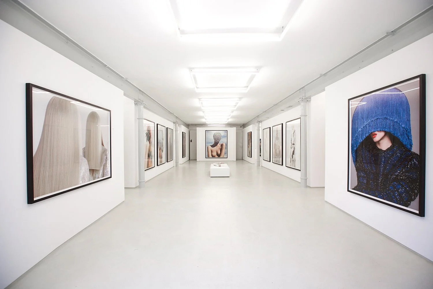 Ein Kunstgalerie-Raum mit weißen Wänden und einer Reihe von großformatigen Fotografien an den Wänden, einige zeigen Frauen in künstlerischen Posen und Verkleidungen, im Mittelpunkt ein Bild einer nackten Frau mit einer Kopfbedeckung aus Bällen.