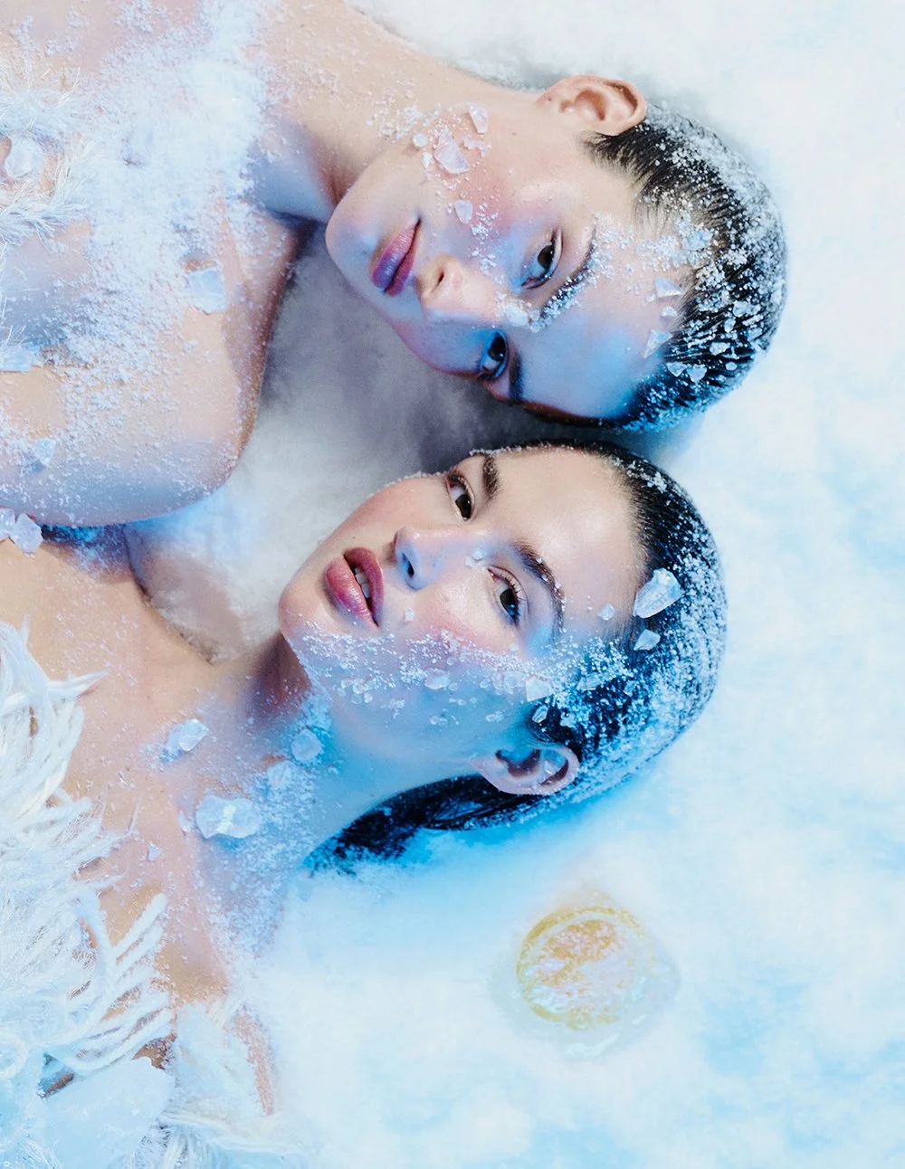 Zwei Frauen liegen nackt in einem Bad, umgeben von weißem Pulver und Wasser, mit verträumtem Blick und nassem, dunklem Haar.