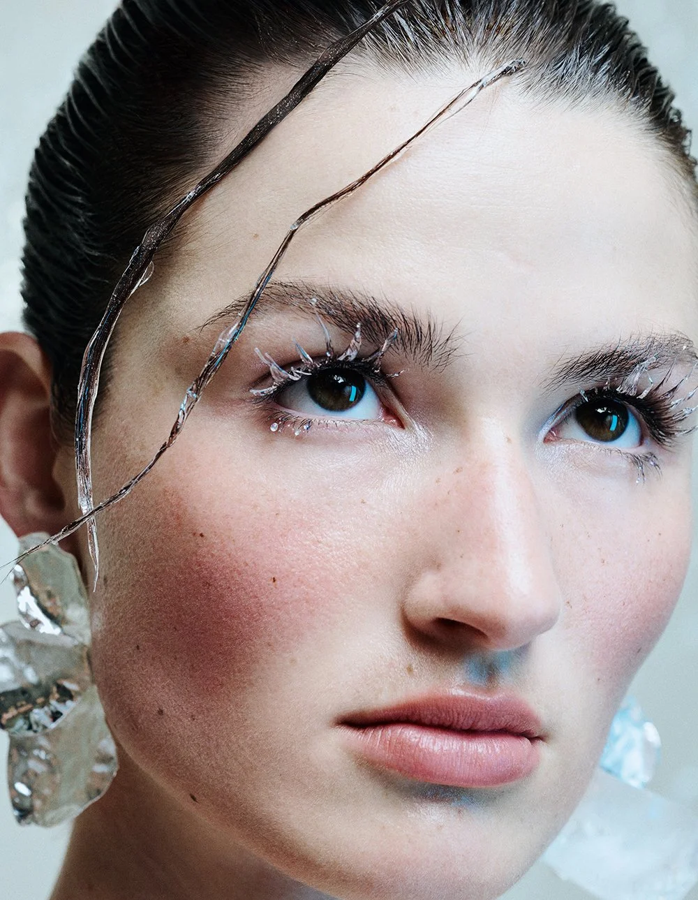 Nahaufnahme eines Gesichts mit ausgefallenen Make-up- und Schmuckdetails, darunter Glas- und Metall-Accessoires, sowie kunstvoll gestaltetem Augen-Make-up.