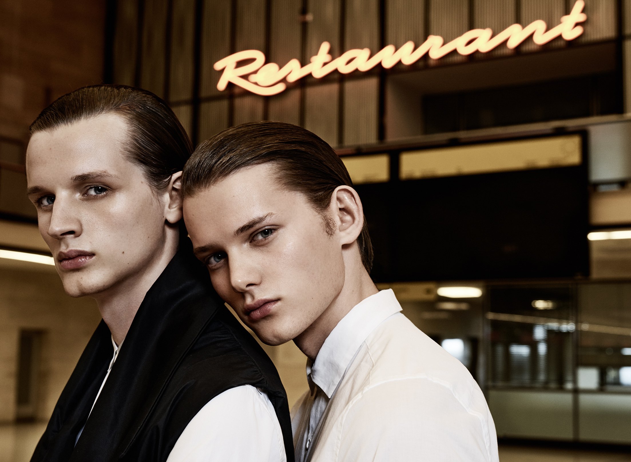 Zwei junge Männer mit glatten, schulterlangen Haaren und blauer Augen stehen nah beieinander vor einem Restaurant, das im Hintergrund leuchtend ist.