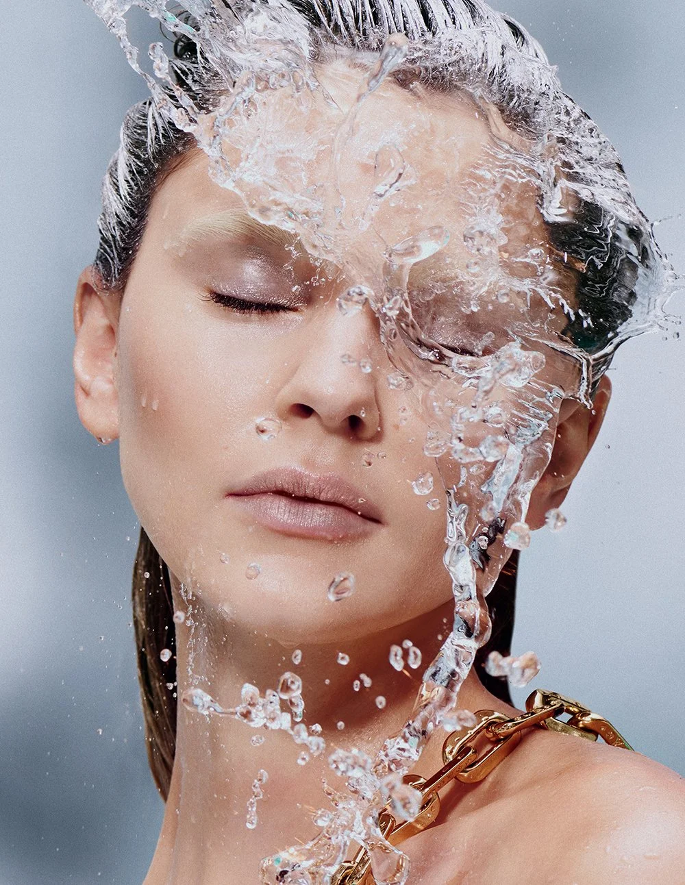 Eine Frau, deren Gesicht mit Wasser bedeckt ist, mit geschlossenen Augen, beim Wasserspiel. Sie trägt einen goldenen Halsketten-Accessoire.