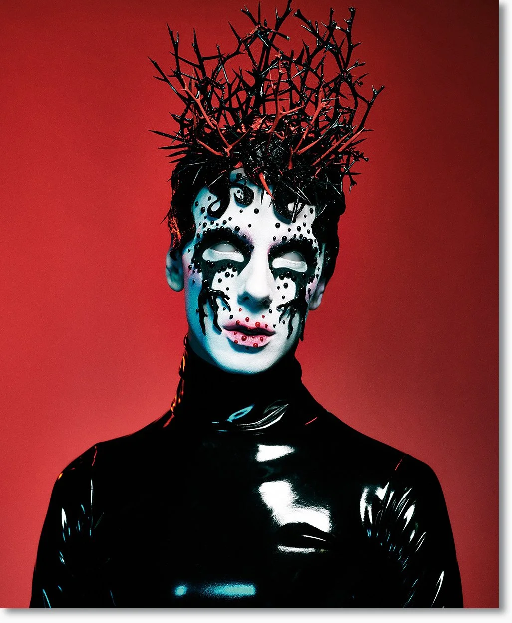 Person mit aufwändiger Schwarz-Weiß-Make-up und roten Akzenten, trägt eine schwarze, glänzende Kleidung, mit einem Koronakranz aus roten Dornen auf dem Kopf, vor rotem Hintergrund.