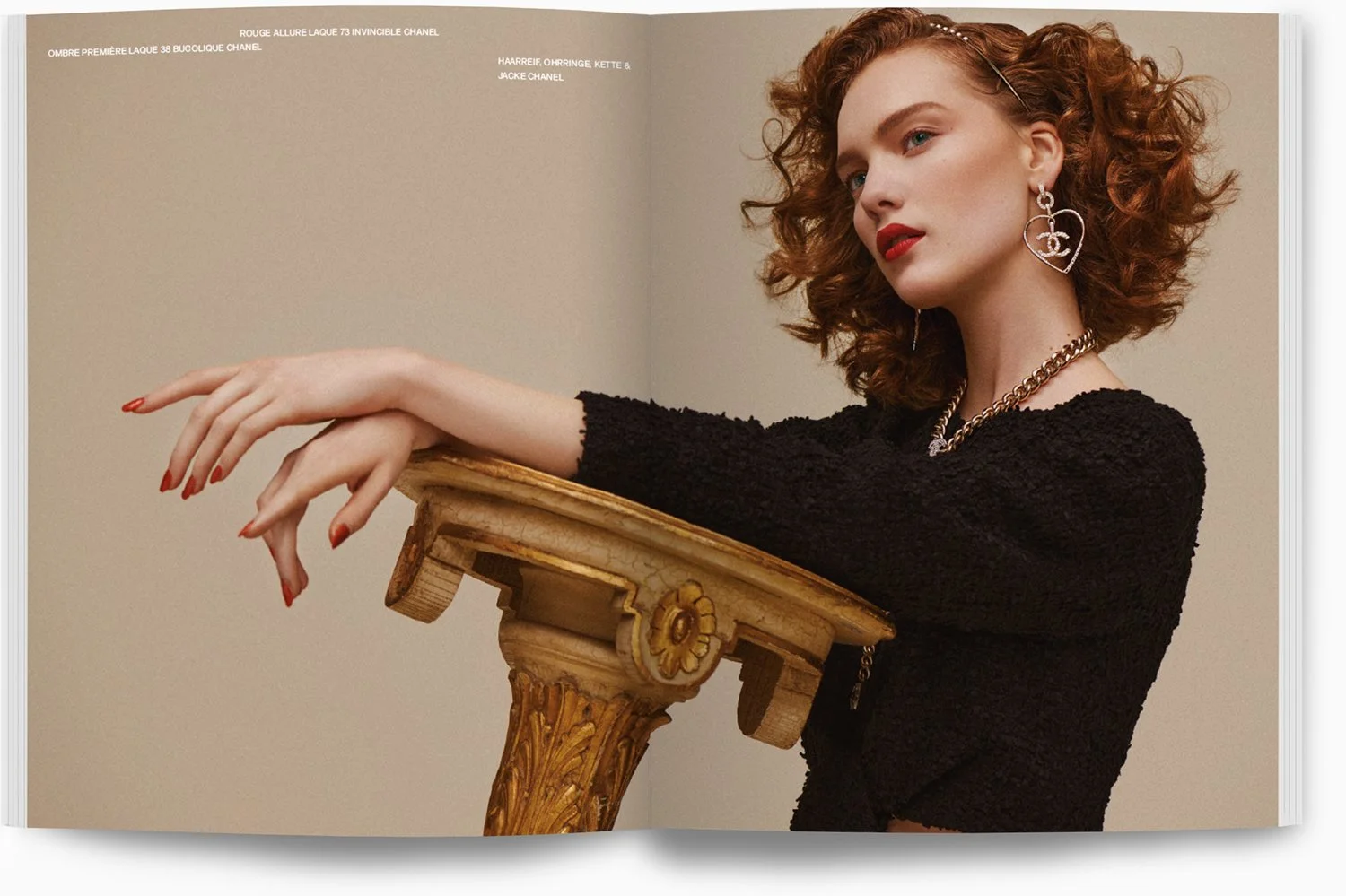 Eine junge Frau mit lockigem roten Haar, die auf einem goldfarbenen, antik aussehenden Tisch lehnt. Sie trägt große Herz-Ohrringe, eine Silberkette und ein schwarzes Oberteil. Der Hintergrund ist neutral beige.