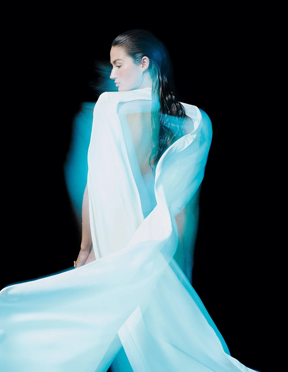 Frau mit nassem Haar, in einem weißen Kleid, das sich im Wind bewegt, vor schwarzem Hintergrund.