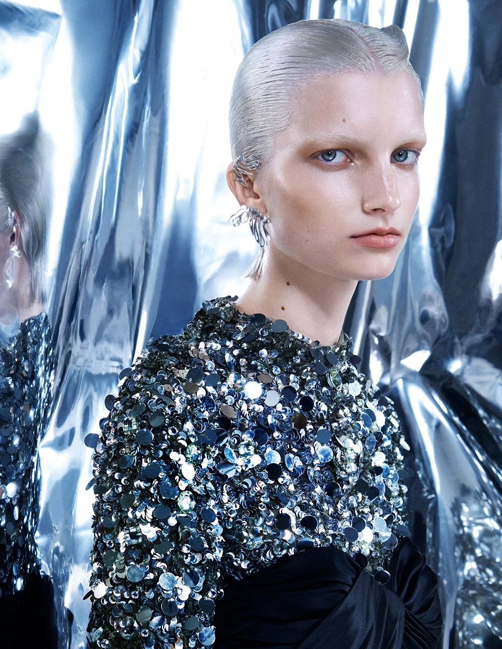 Ein Model mit blondem, nassen Haar und blauen Augen trägt ein schwarzes Kleid mit silbernen Pailletten und steht vor einer metallic silbernen Kulisse.