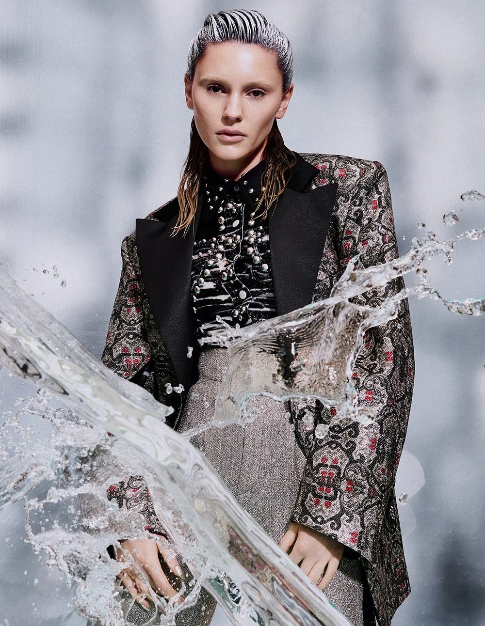 Frau in hochmodischer Kleidung steht vor einem neutralen Hintergrund, Wasser spritzt dynamisch im Vordergrund.