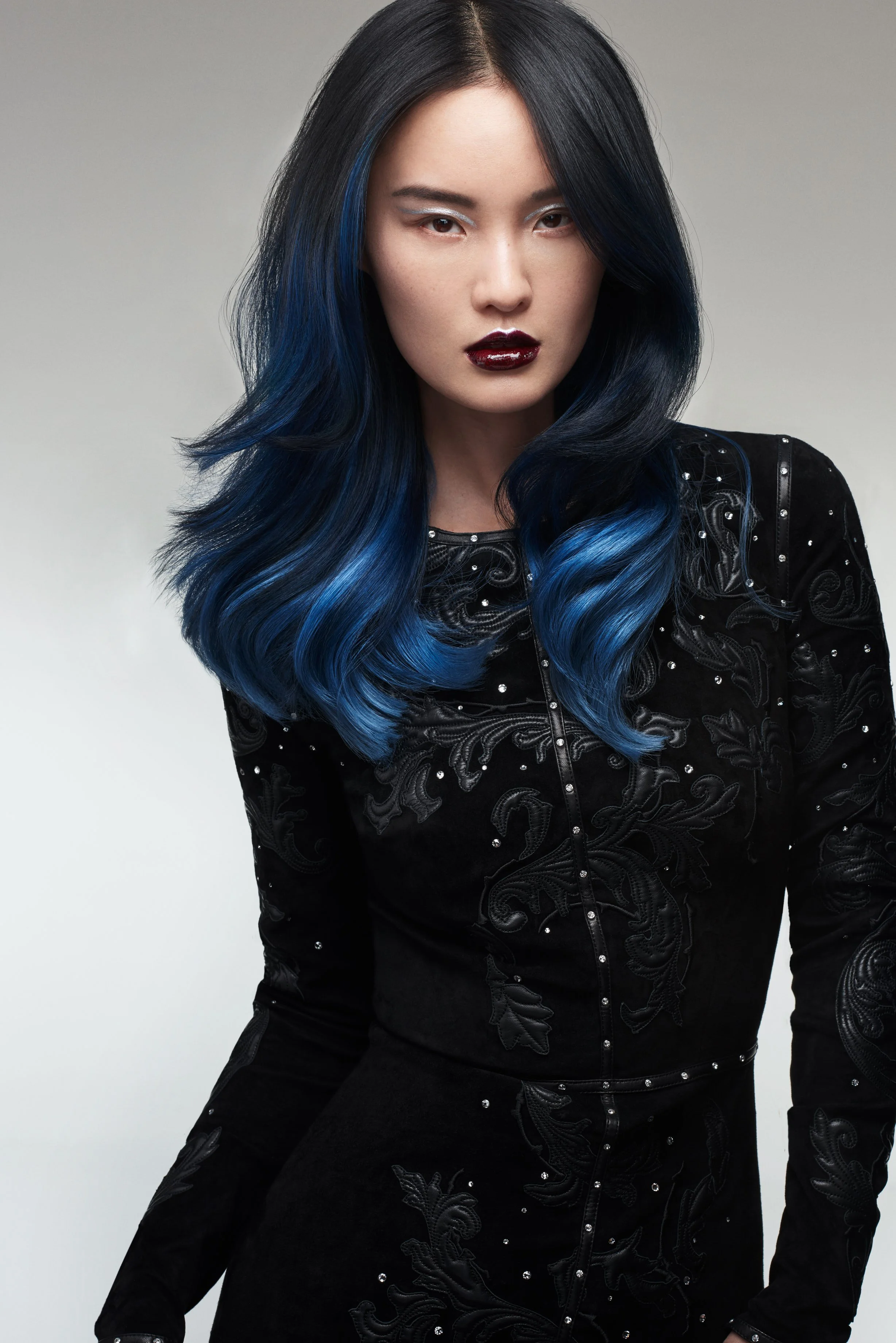 Junge Frau mit schwarzen Haaren, blauen Hervorhebungen, dunklem Lippenstift, schwarzem Kleid mit Stickereien und Glitzern.