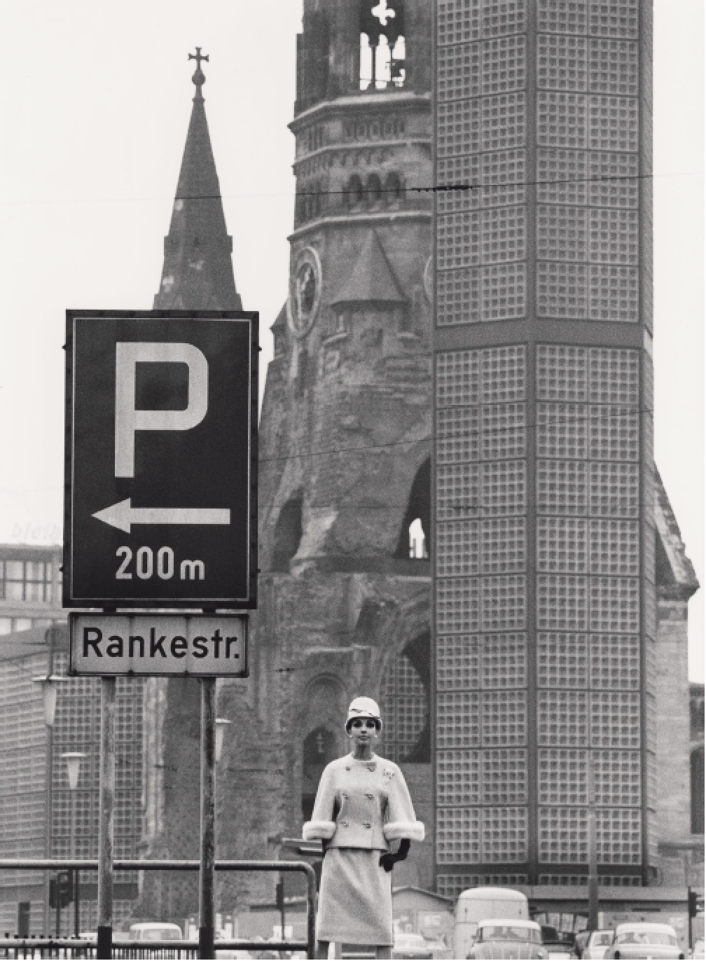 Eine Frau in eleganter Kleidung steht vor einem historischen Gebäude mit einem Kirchturm im Hintergrund. Es ist eine schwarz-weiße Aufnahme.