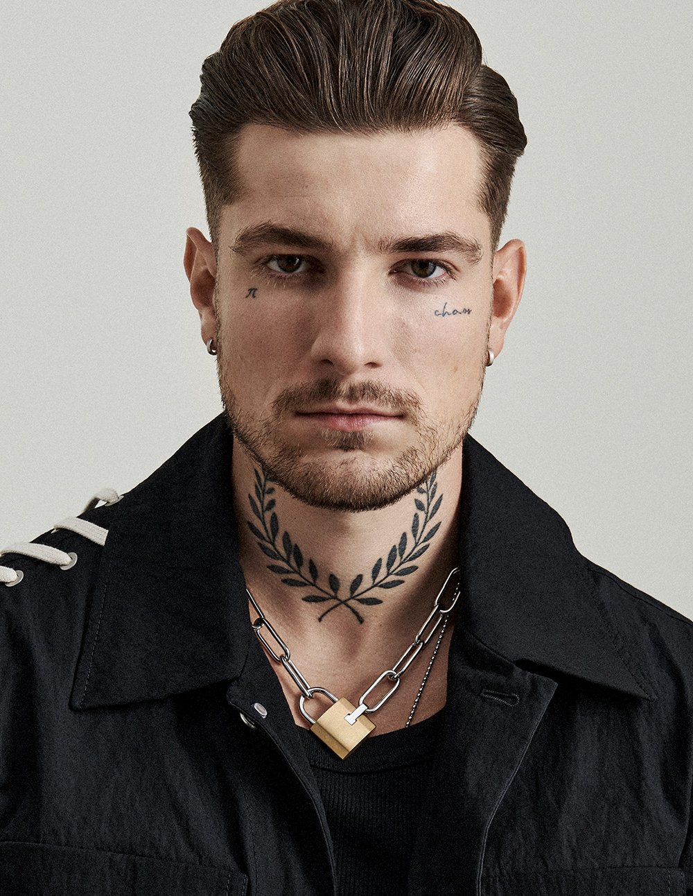 Ein junger Mann mit kurzen braunen Haaren, Starkem Bart, Tattoos im Gesicht und am Hals, trägt eine schwarze Jacke und eine große Kette mit Vorhängeschloss.