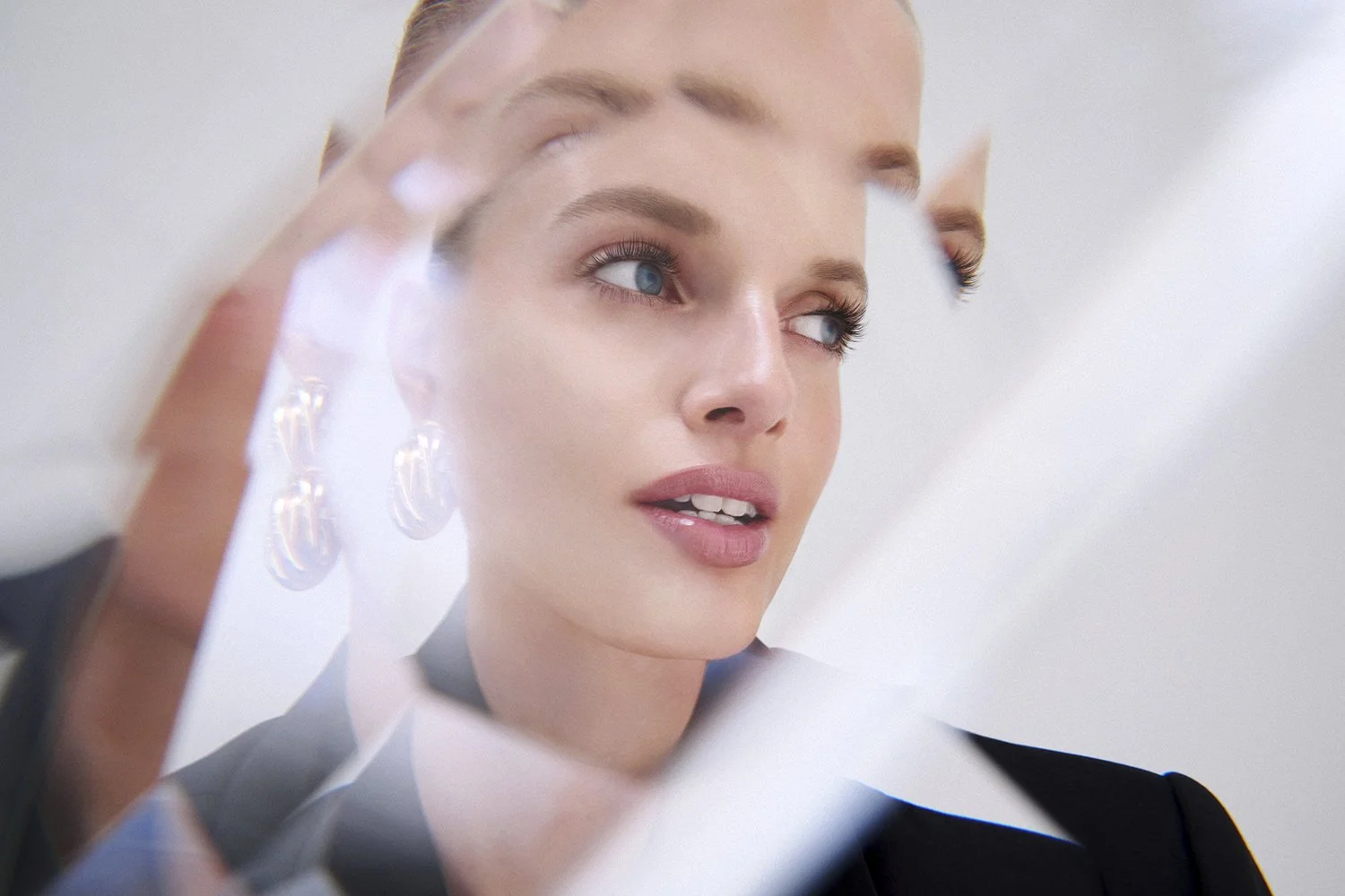 Close-up of a young woman looking at her reflection in a hand mirror, with blurred earrings and light-colored background.