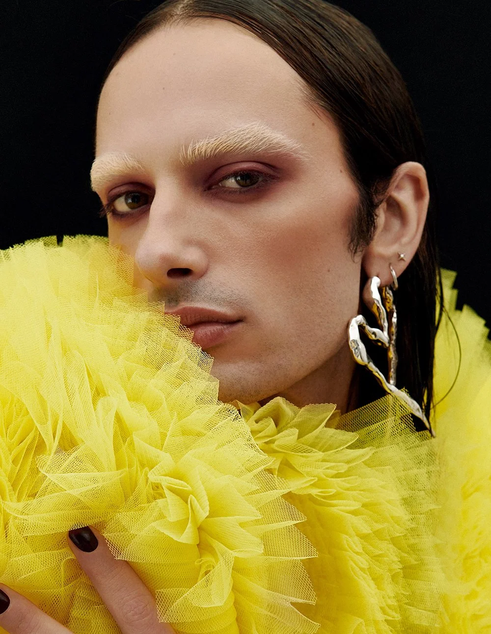 Porträt eines Mannes mit hellen Augen, blonden Augenbrauen, dunklen Haaren und mehreren Ohrringen, vor einem schwarzen Hintergrund, trägt ein auffälliges gelbes Kleid mit großen Tüllblumen.