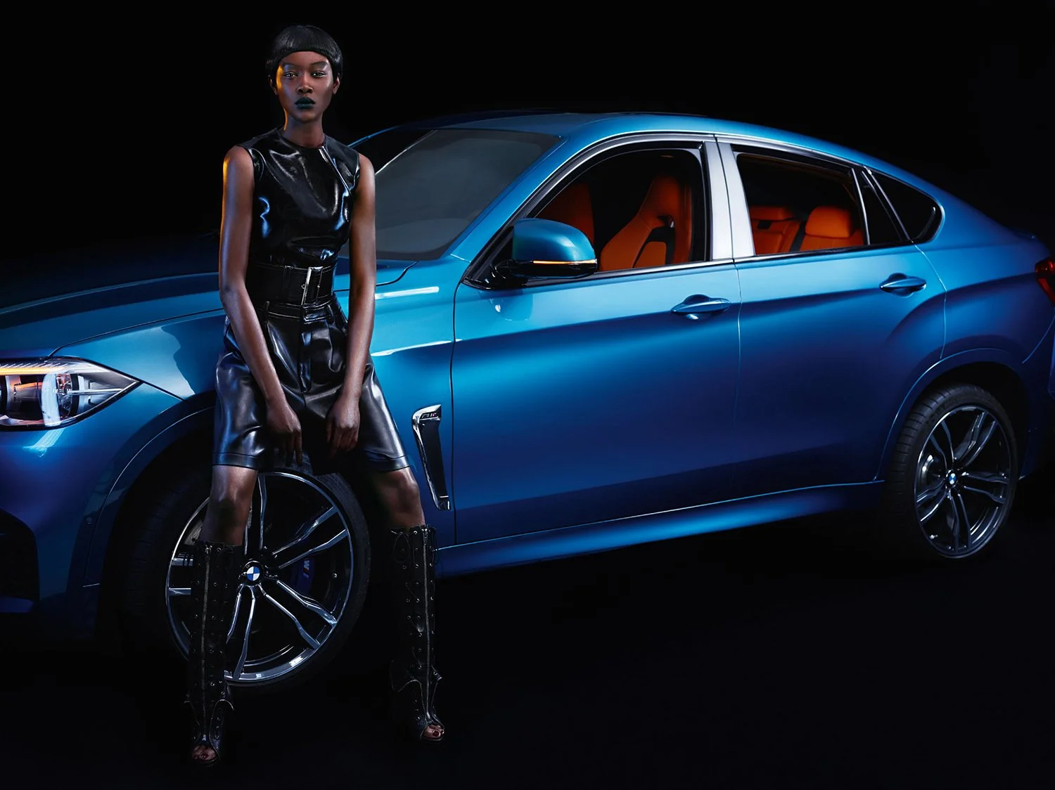 A woman dressed in black leather outfit and boots standing next to a blue luxury car with orange interior, against a black background.