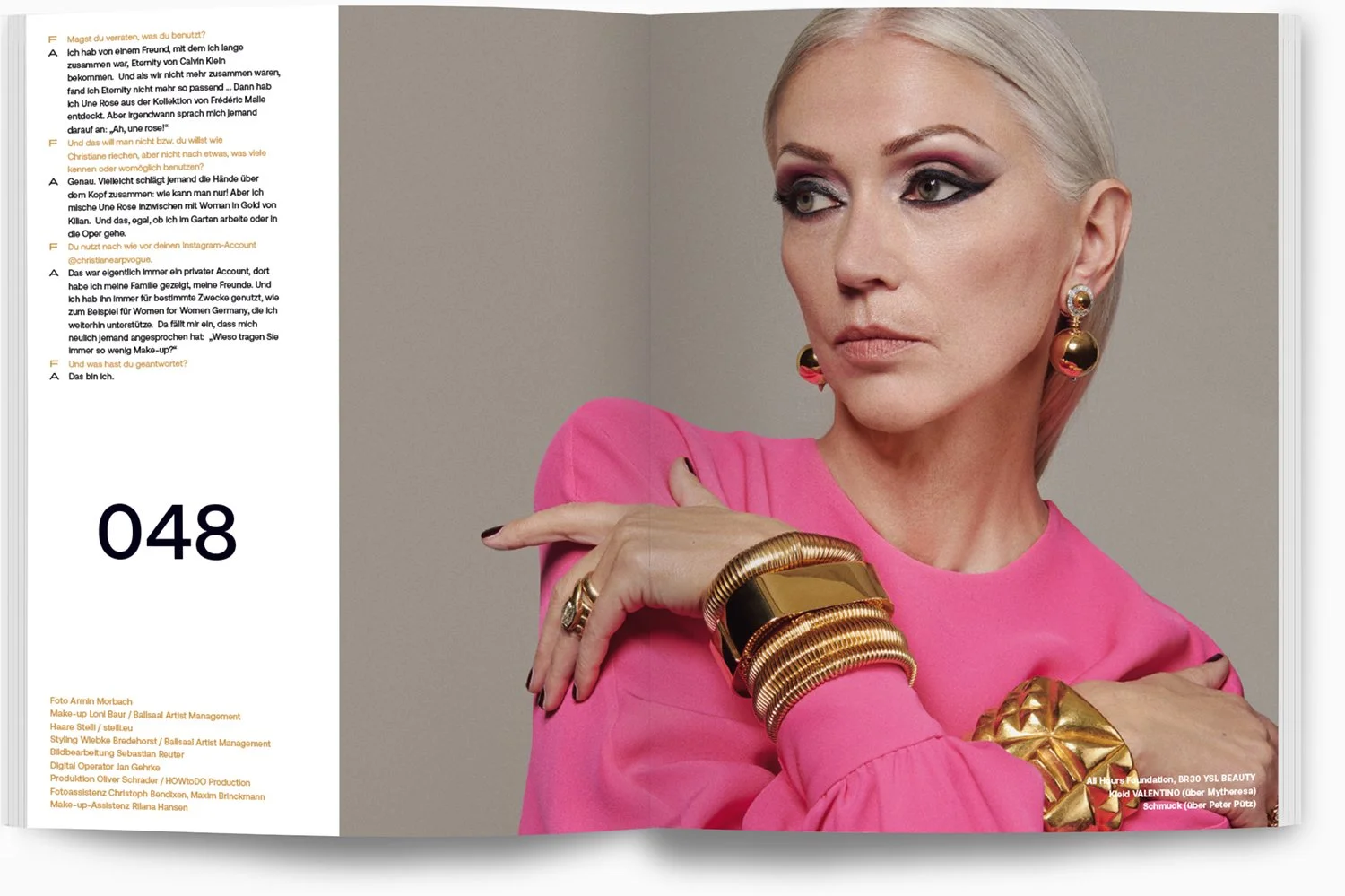 Eine Frau mit platinumblonden Haaren trägt große goldene und pinke Ohrringe, mehrere goldene Armbänder und einen goldenen Ring, während sie ein pinkes Oberteil trägt. Sie hat aufwändiges Make-up mit dunklen Lidschatten und Lippen.