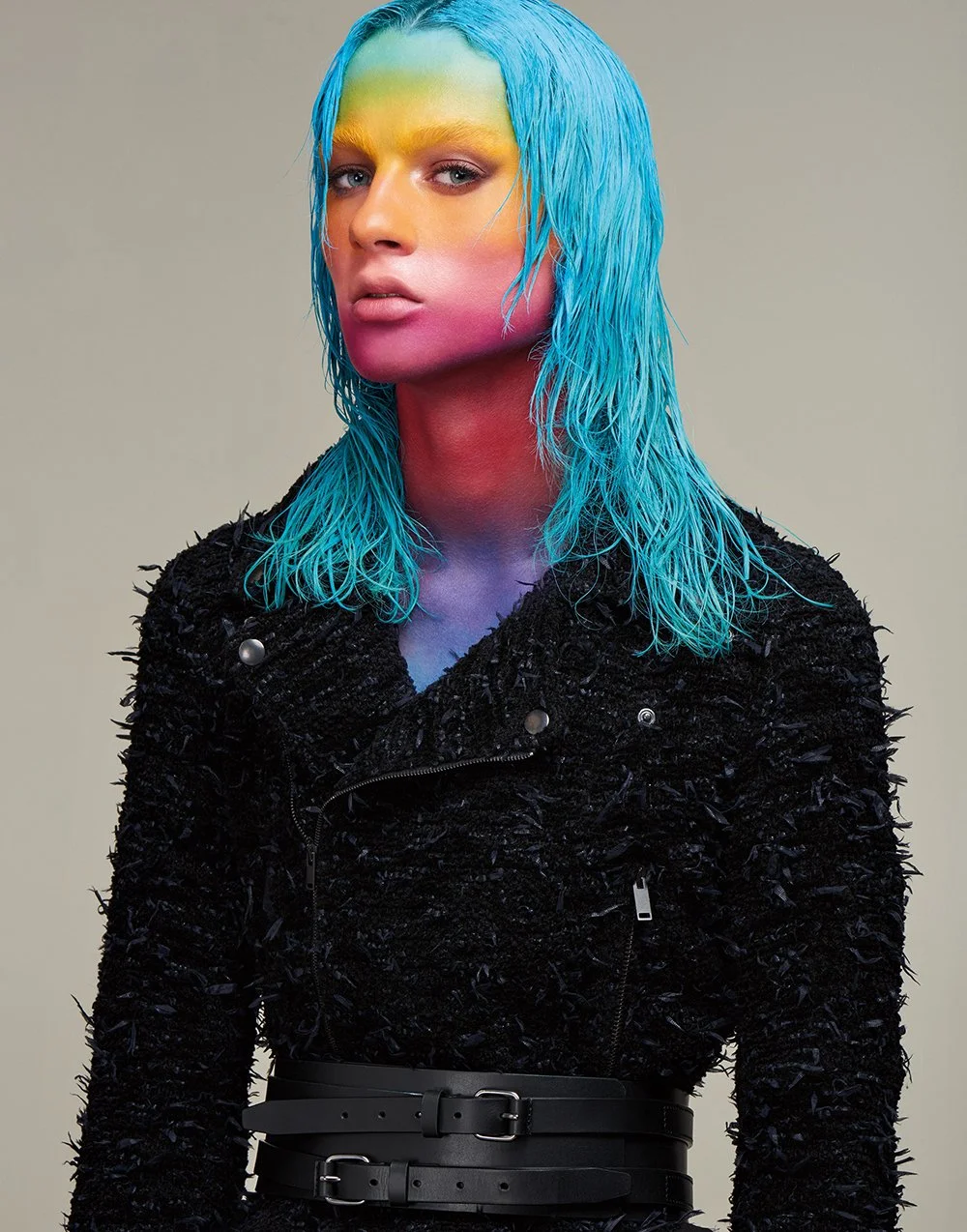 Frau mit blauen, nassen Haaren und Farbmuster im Gesicht und Hals, trägt schwarze, strukturierte Jacke und einen breiten schwarzen Gürtel.