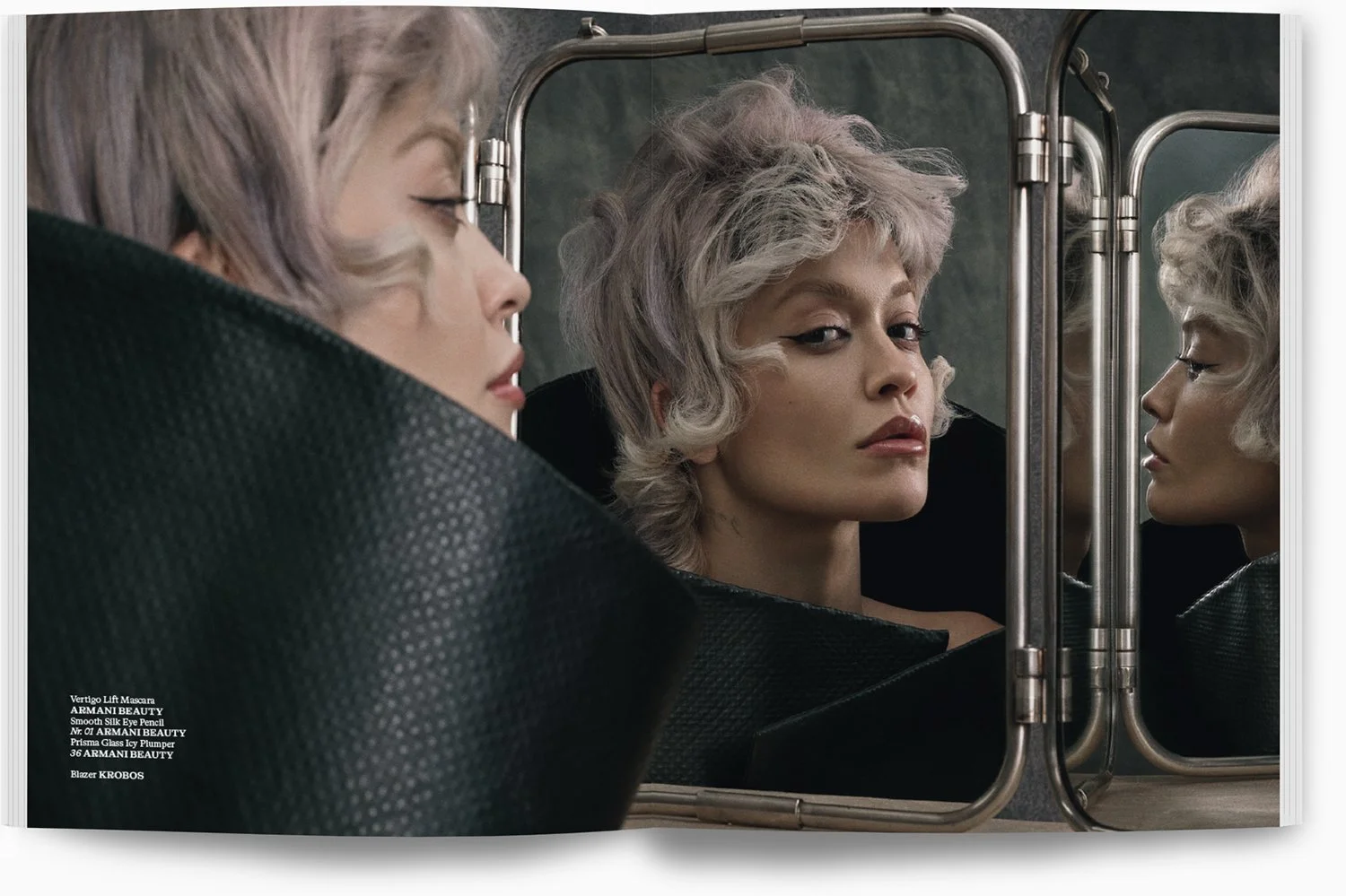 Ein Model mit kurzen, lockigen, hellblonden Haaren blickt in einen Spiegel, wodurch sie von verschiedenen Blickwinkeln sichtbar ist. Das Bild hat eine dunkle, elegante Atmosphäre und betont das Make-up sowie die schwarzen, strukturierten Kleidung des