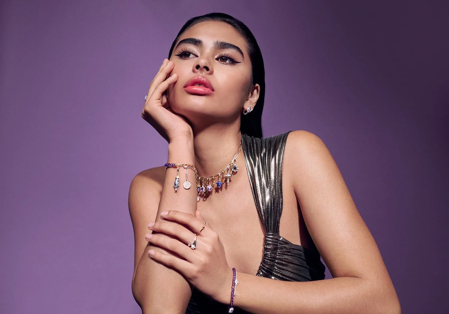 A woman with dark hair and makeup wearing a sleeveless metallic top, adorned with jewelry including necklaces, rings, and bracelets, posed against a purple background.