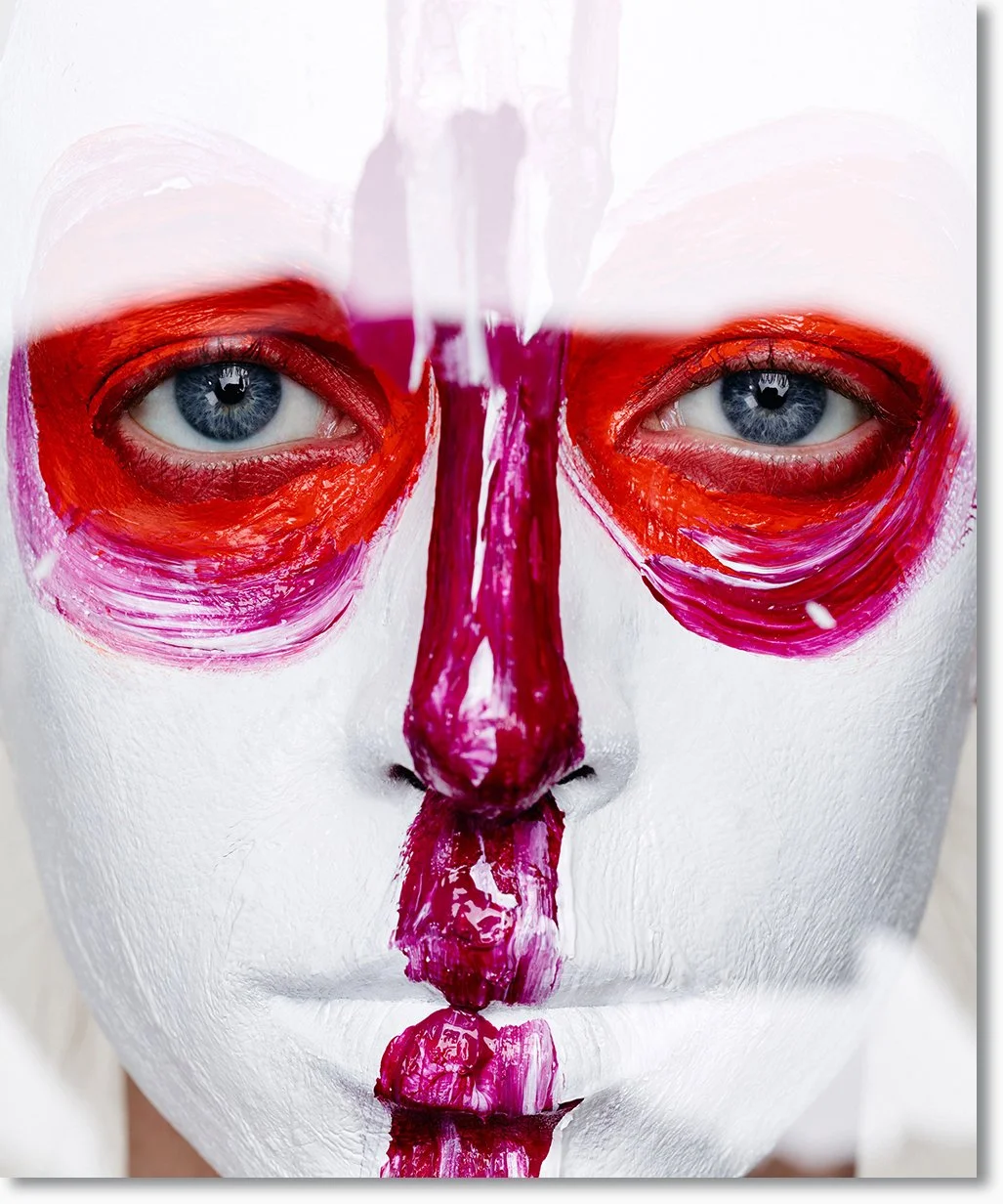 Nahaufnahme eines Gesichts mit weißen Gesichtsbemalung und roten, lila Akzenten um die Augen, die Nase und den Mund, Betonung auf blaue Augen.