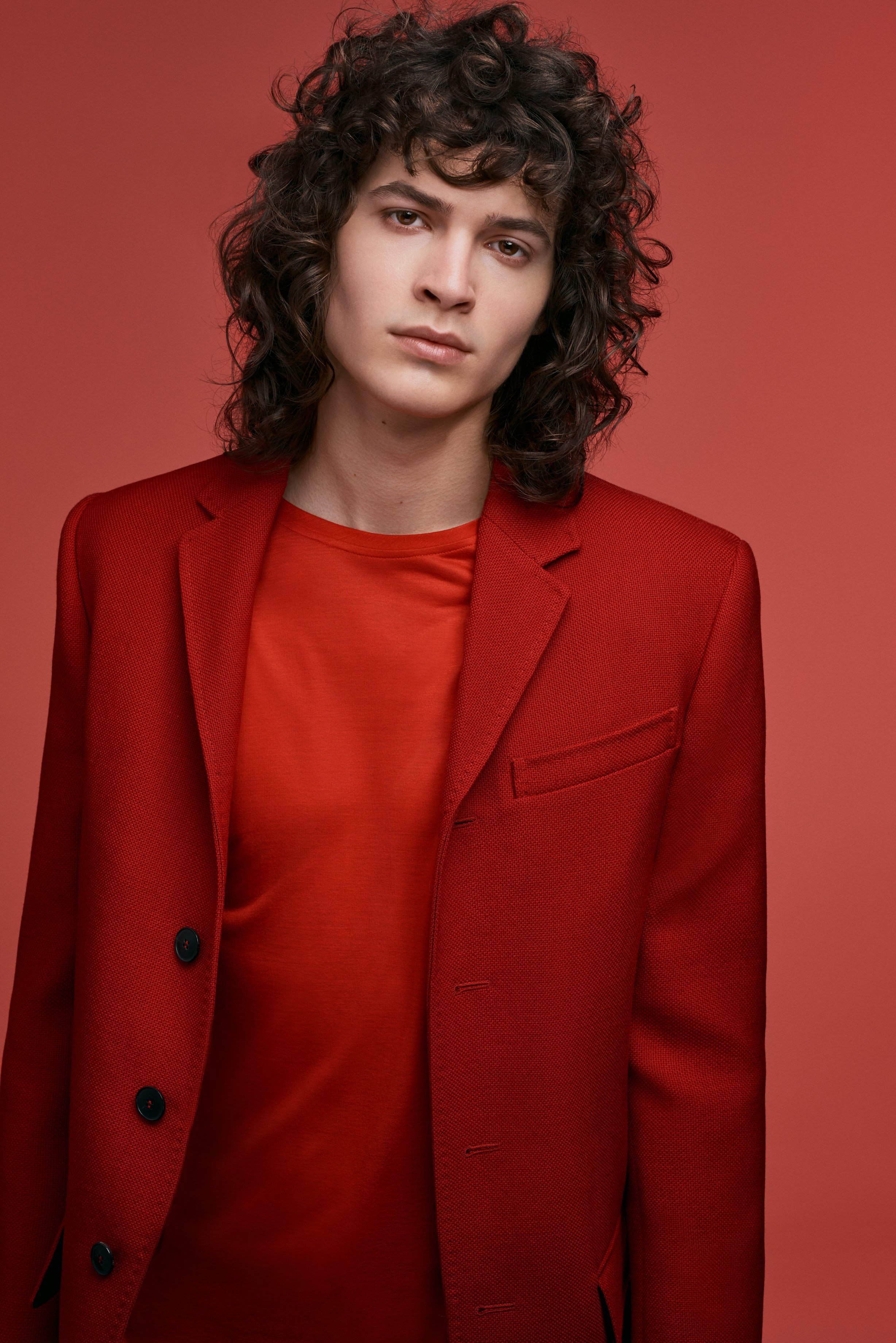 Porträt eines jungen Mannes mit lockigem, dunklem Haar, in rotem Anzug und rotem Shirt vor rotem Hintergrund.