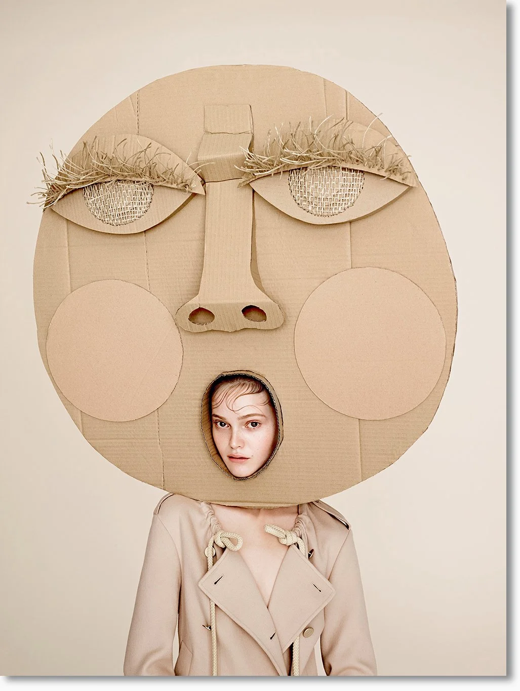 Junge trägt eine große Porträtmaske aus Pappe, die ein Gesicht mit geschlossene Augen, großen Wangen, Nase und Mund mit einem Loch für das Gesicht zeigt.