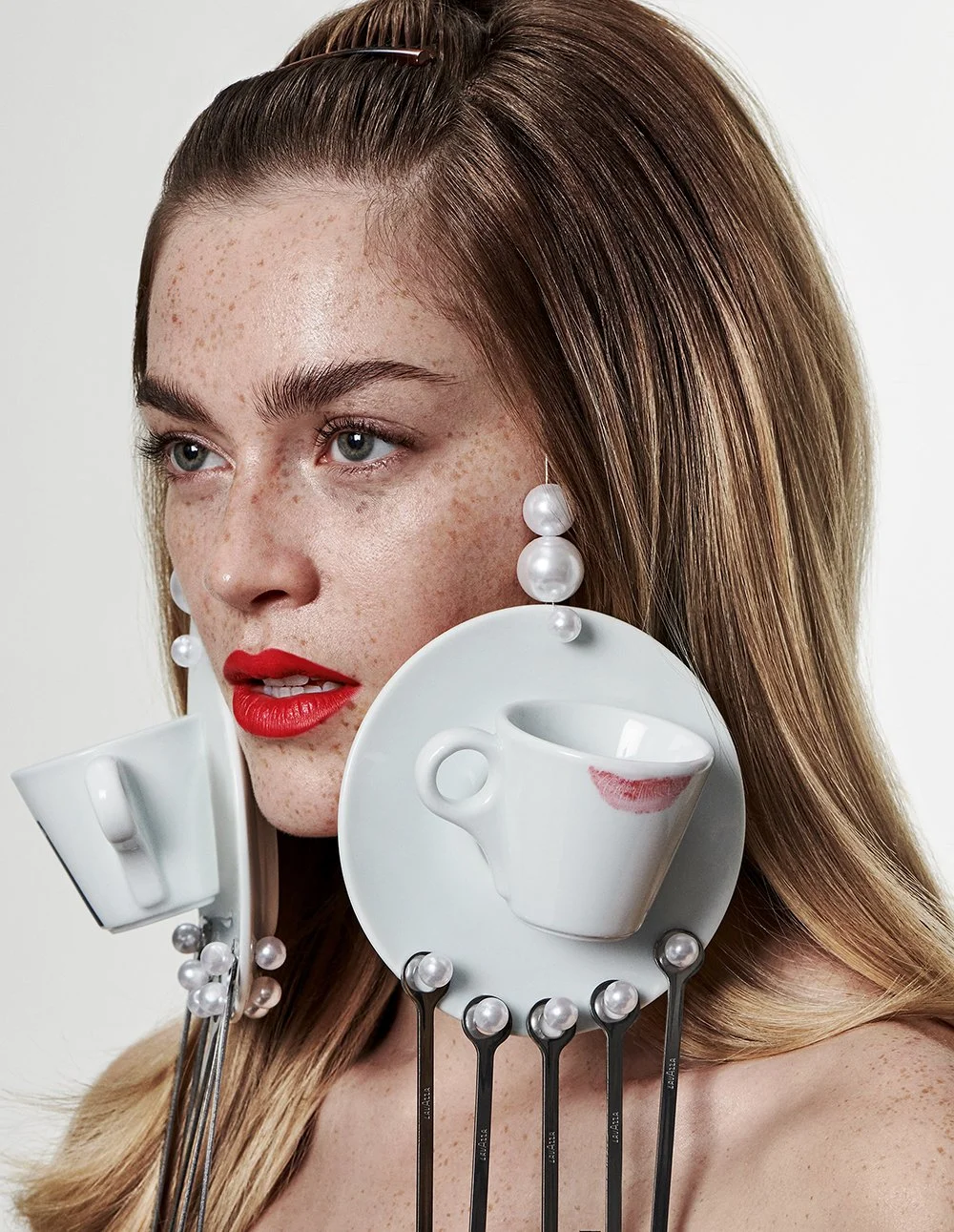 Porträt einer Frau mit rotem Lippenstift und hellbraunen Haaren, umgeben von weißen Porzellan-Tassen und Löffeln mit Perlen, die auf einer weißen Platte befestigt sind.