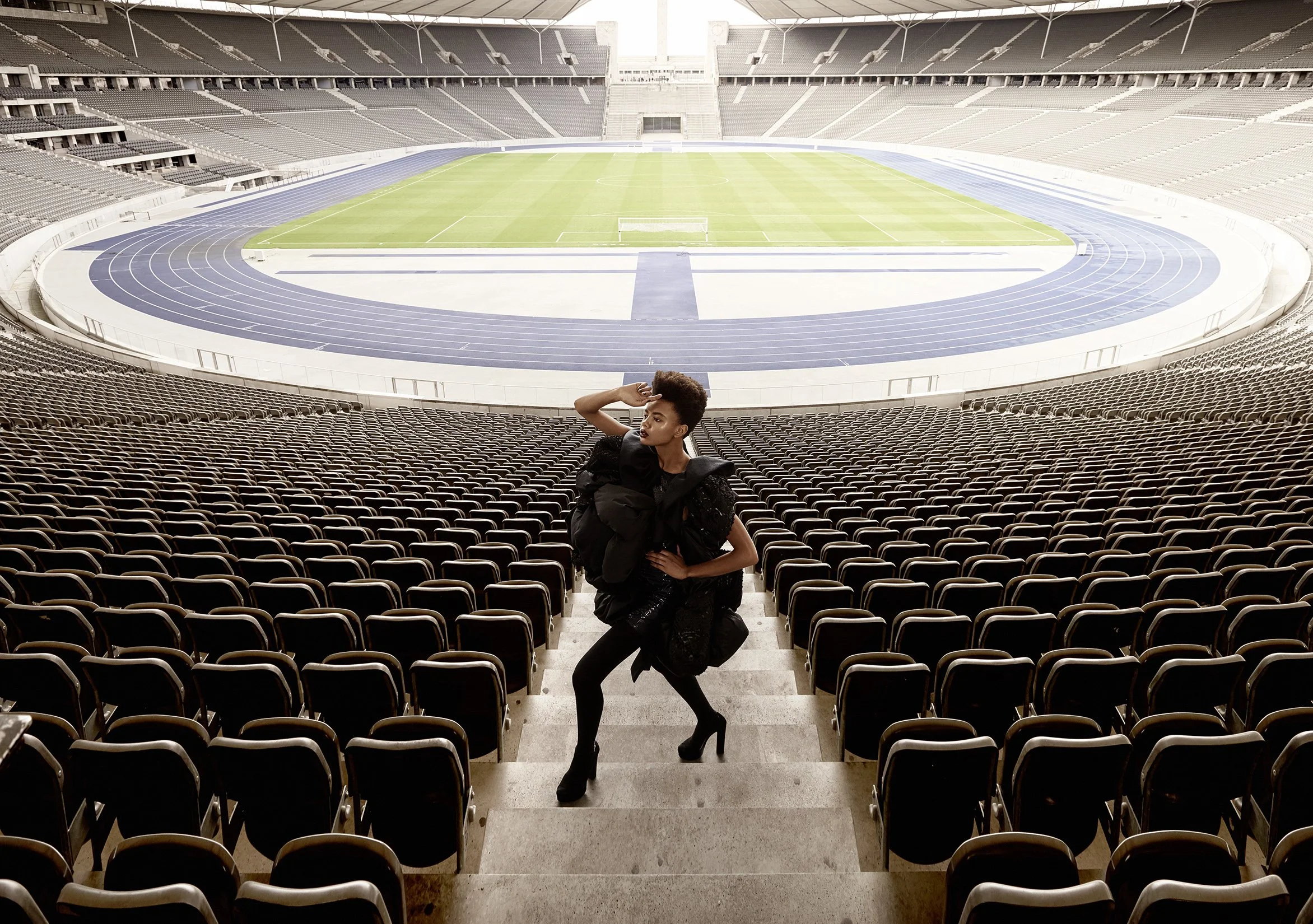 Eine Frau in Hochhackschuhen und mit vielen schwarzen Taschen steht in einem leeren Stadion, Blick auf den leeren Fußballplatz und die Laufbahn.