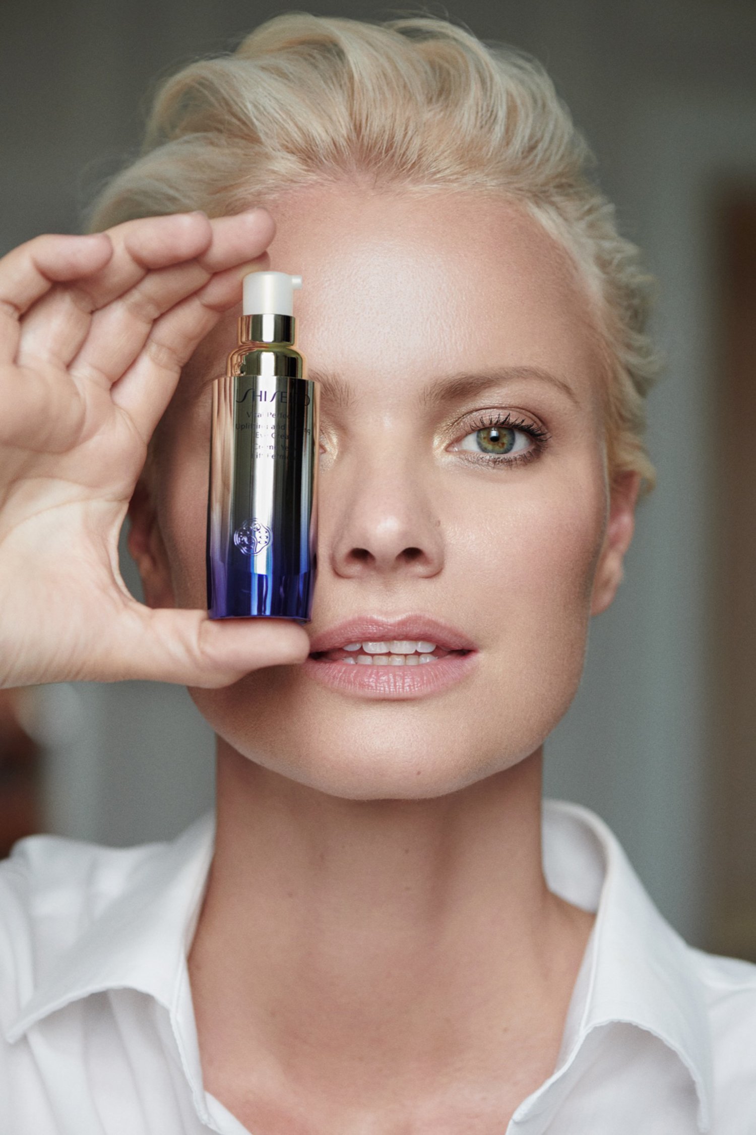 A woman holding a skincare product in front of her face, showing only her head and shoulders, with a neutral background.
