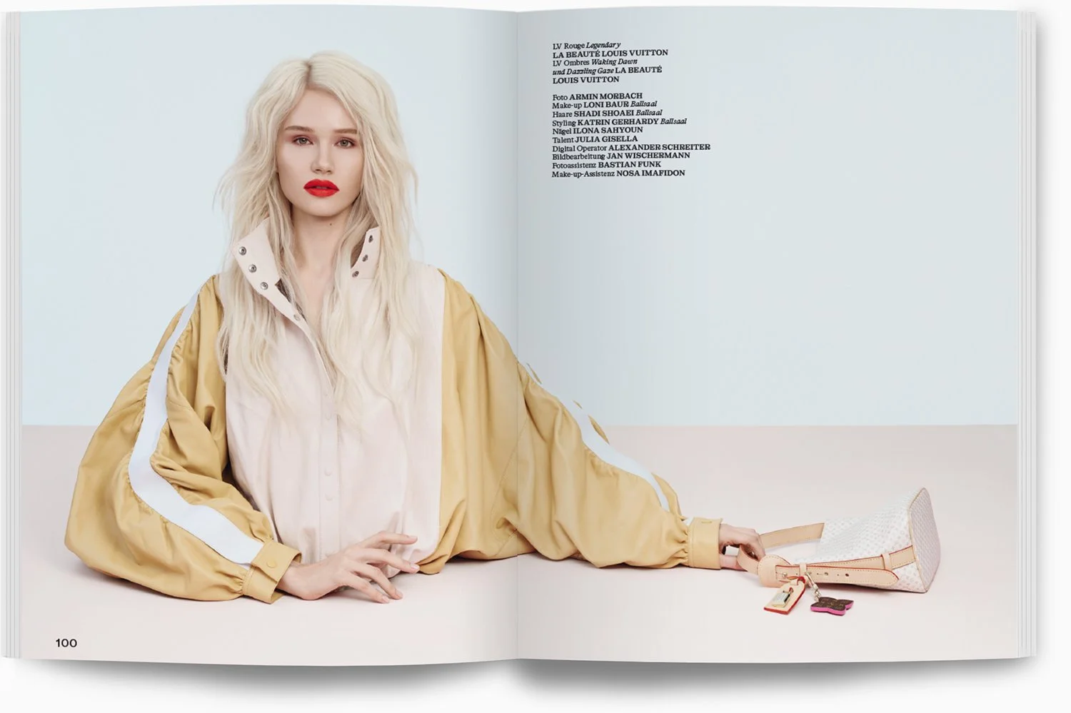 Eine blonde Frau liegt auf dem Boden in einem Modefoto. Sie trägt ein beige-weißes Oberteil und eine beige Jacke, hält eine kleine Tasche mit Schlüsselanhänger, im Hintergrund ein offenes Magazin mit Text.