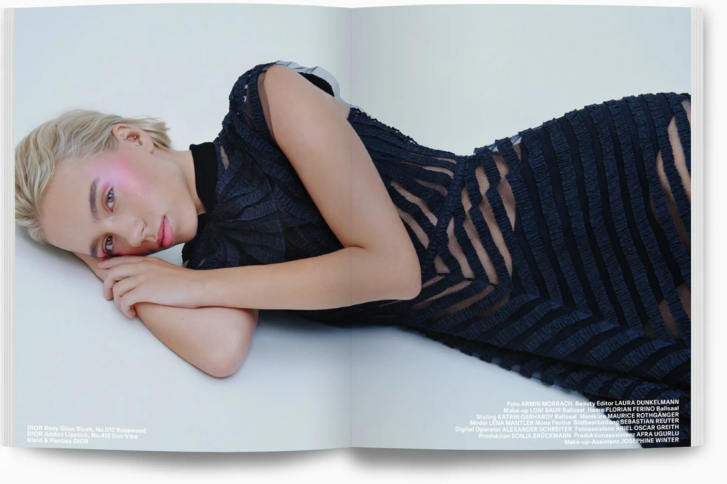 Frau mit blonder kurzen Haaren in einem schwarzen, streifenförmigen Kleid liegt auf einem weißen Boden, blickt in die Kamera, mit leichtem Lächeln und natürlichem Make-up.