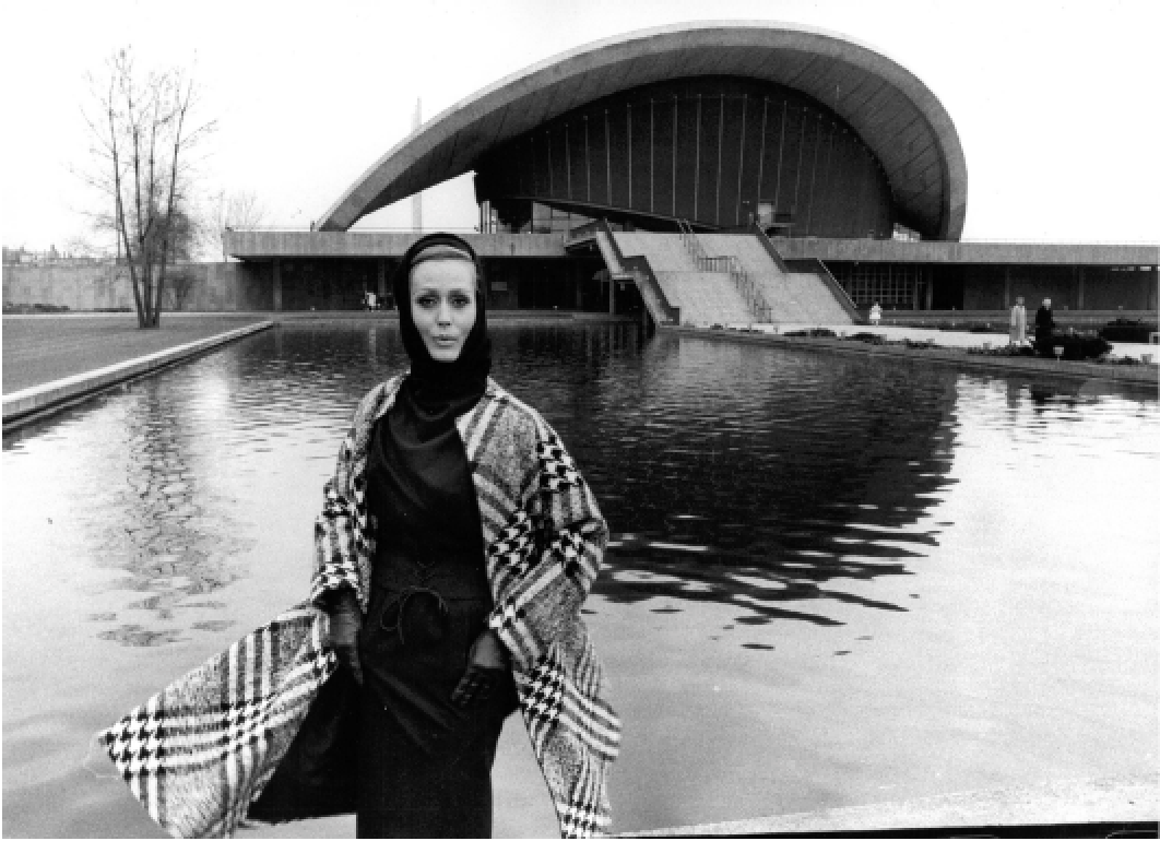 Eine Frau steht vor einem Wasserbecken mit einem modernen, halbrunden Gebäude im Hintergrund, vermutlich ein Theater oder Konzertsaal, in Schwarz-Weiß-Fotografie.