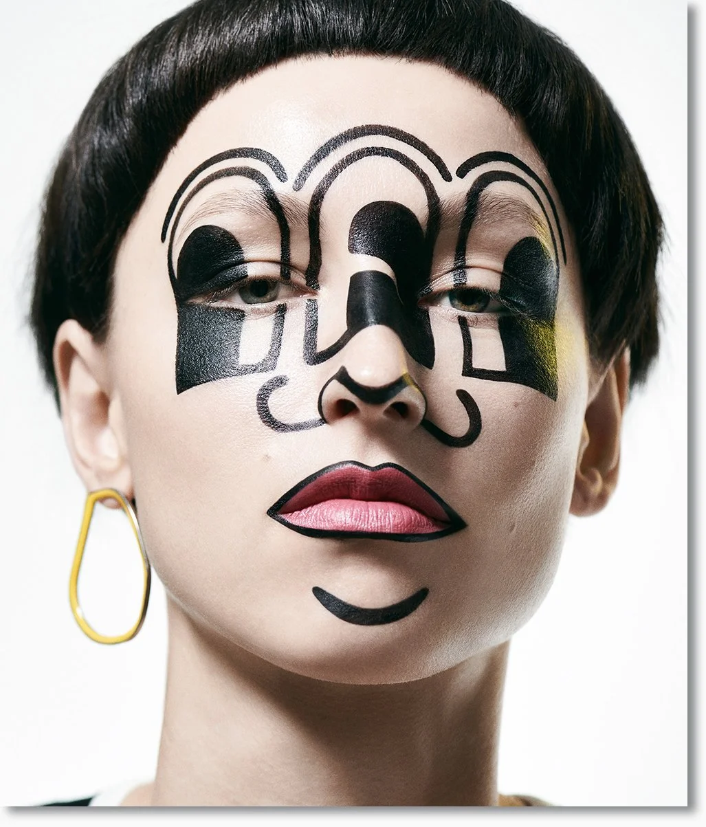 Porträt einer Frau mit Gesichtsbemalung im Stil eines Schädels, schwarzer Lippenstift, dunkelbraune kurze Frisur, lila Augen, gelber Ohrring, weißer Hintergrund.