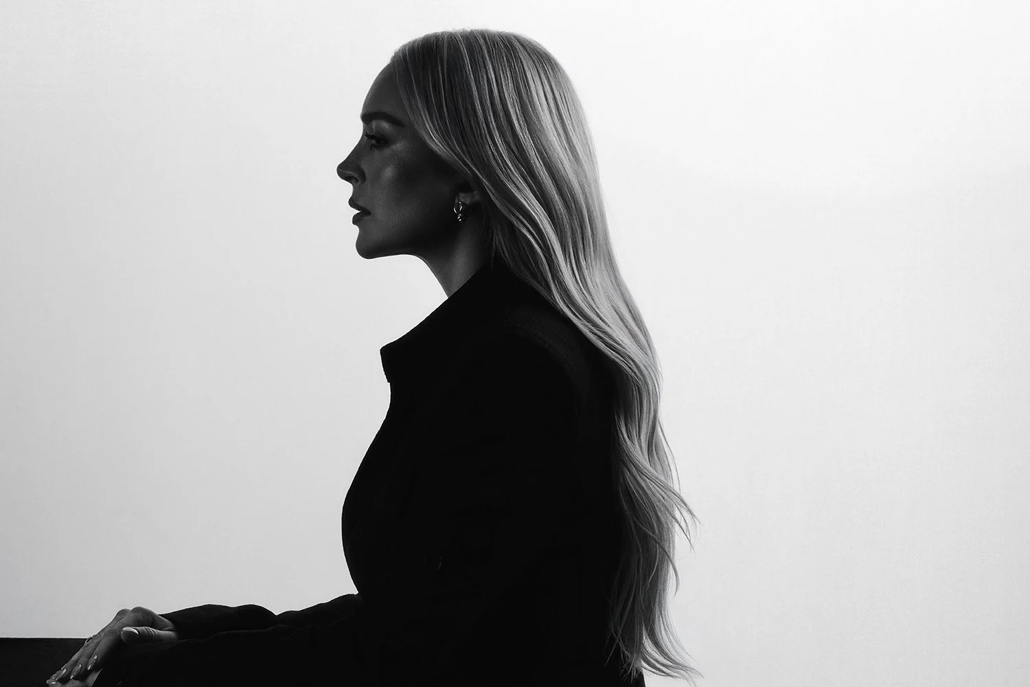 Schwarz-weiß Porträt einer sitzenden Frau mit langen, welligen Haaren, seitlich betrachtet vor einem hellen Hintergrund.