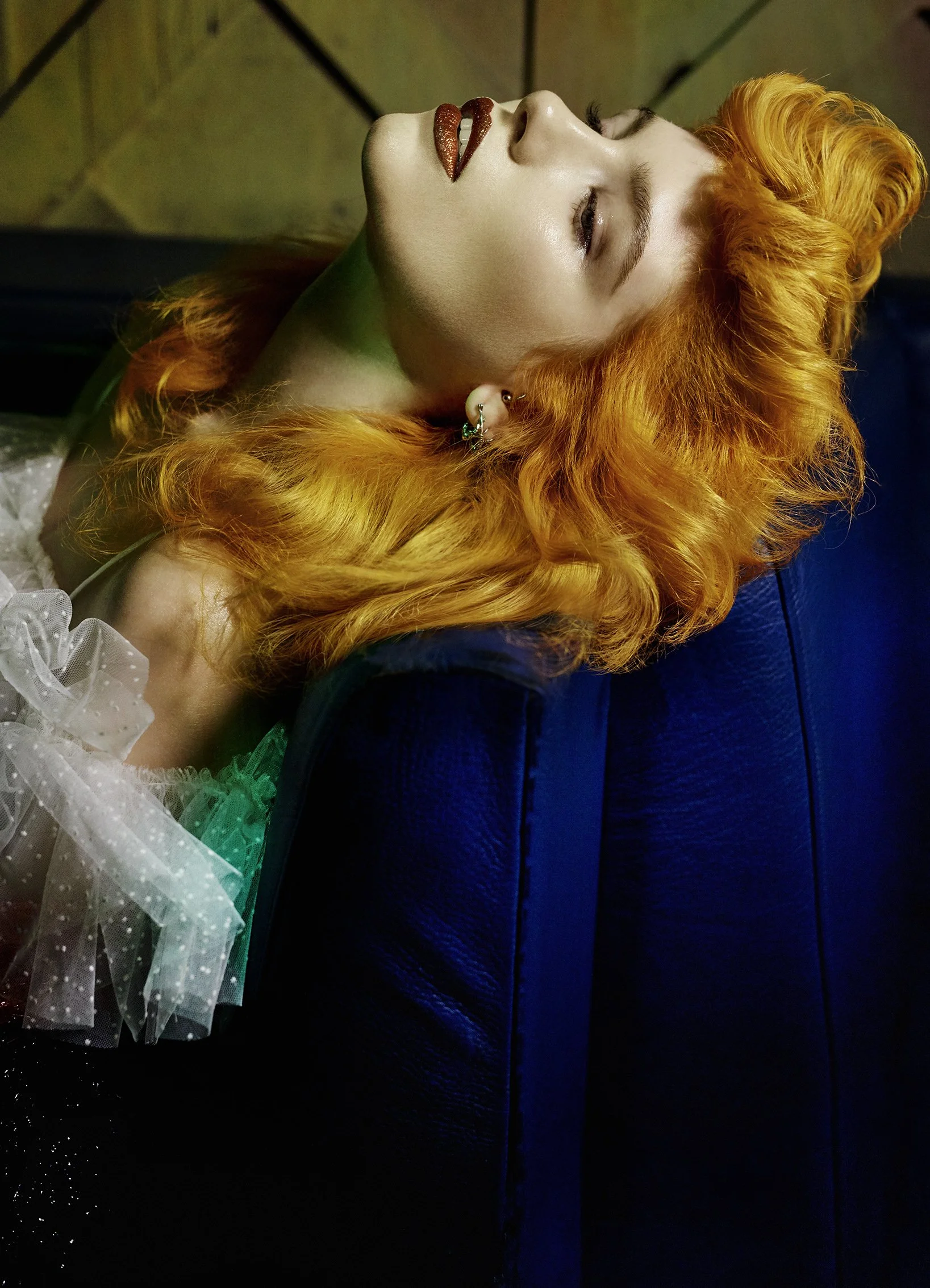 Frau mit orange-lockigem Haar liegt auf einer blauen Couch, Blick nach oben, mit dunklem Lippenstift und mehreren Ohrringen, trägt ein weißes, gepunktetes Tüllkleid.