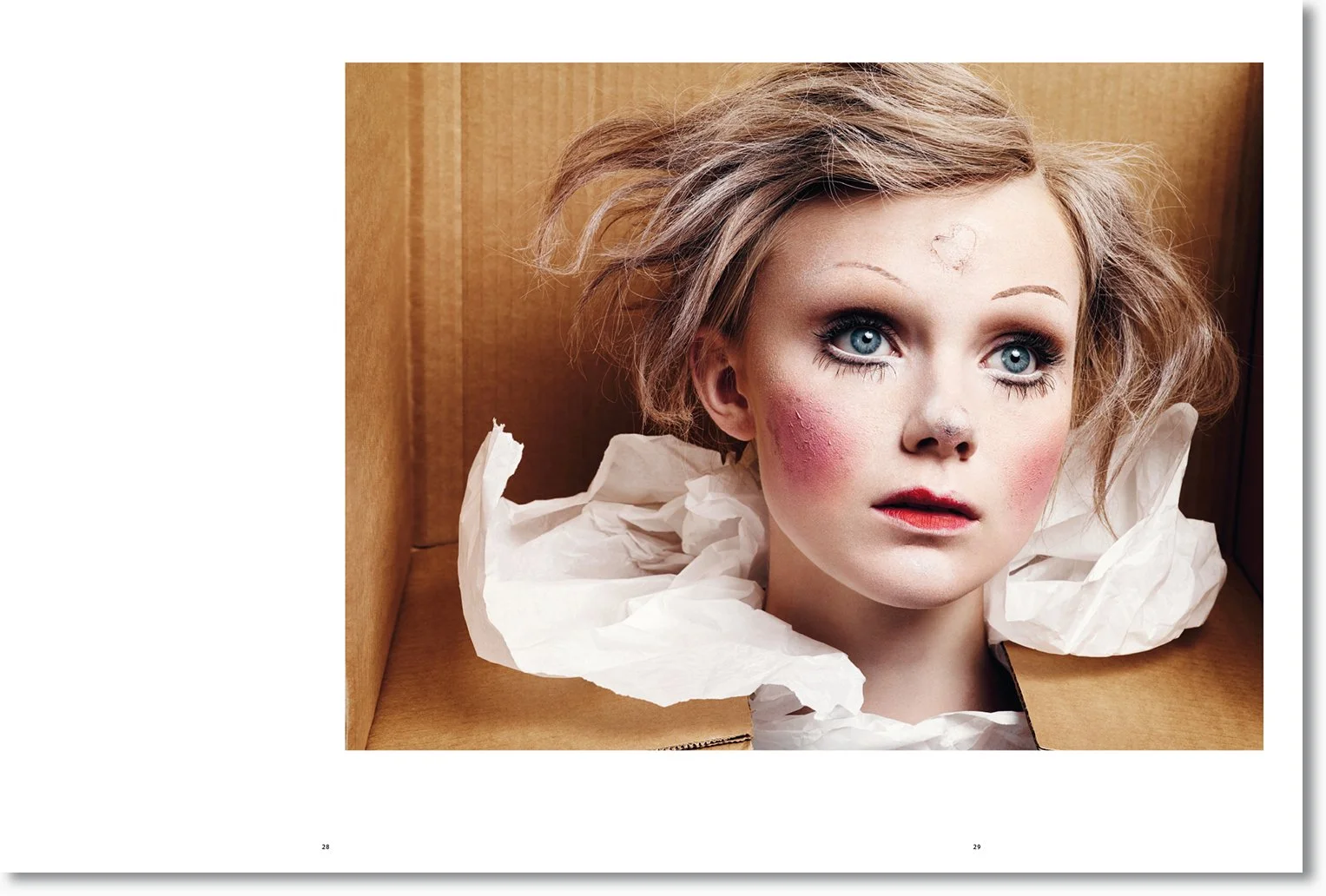 Nahaufnahme eines Mädchens mit blauen Augen, blonder lockiger Haare, rotem Lippenstift und Make-up, das in einer Kartonschachtel mit Papier eingeschlossen ist.