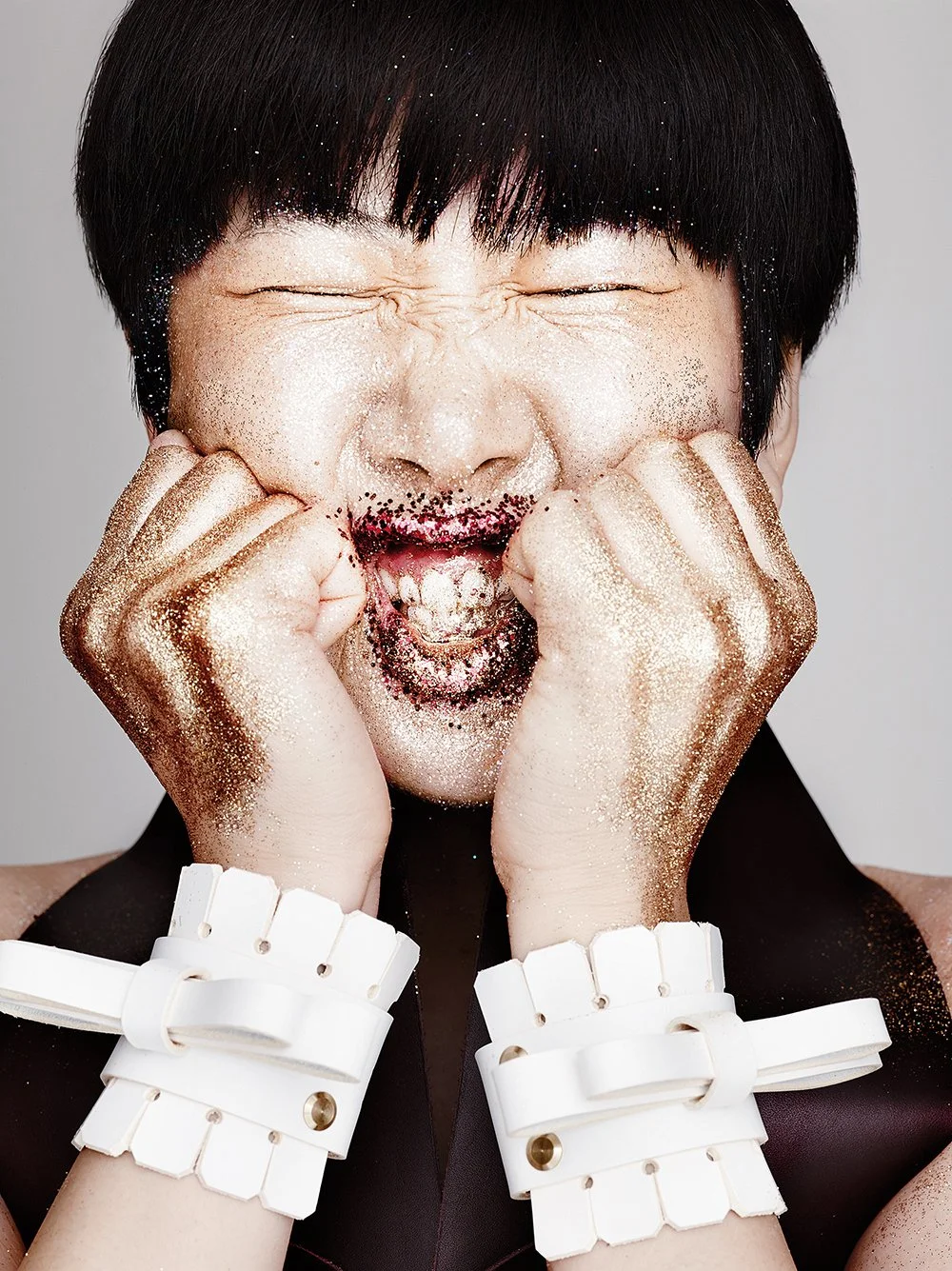 Eine Person mit schwarzhaarigem Bob, die vor einem grauen Hintergrund mit Gold- und schwarzen Glitter-Händeschmuck auf beiden Händen, die die Wangen berühren, eine emotional intensive Szene zeigt.