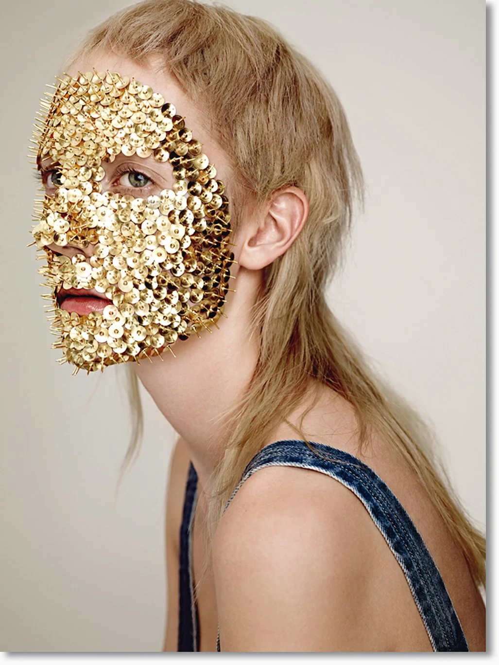 Person mit goldener, ornamentaler Maske, die das Gesicht bedeckt, nur ein Auge sichtbar. Blonde, lockige Haare und rotes Top.