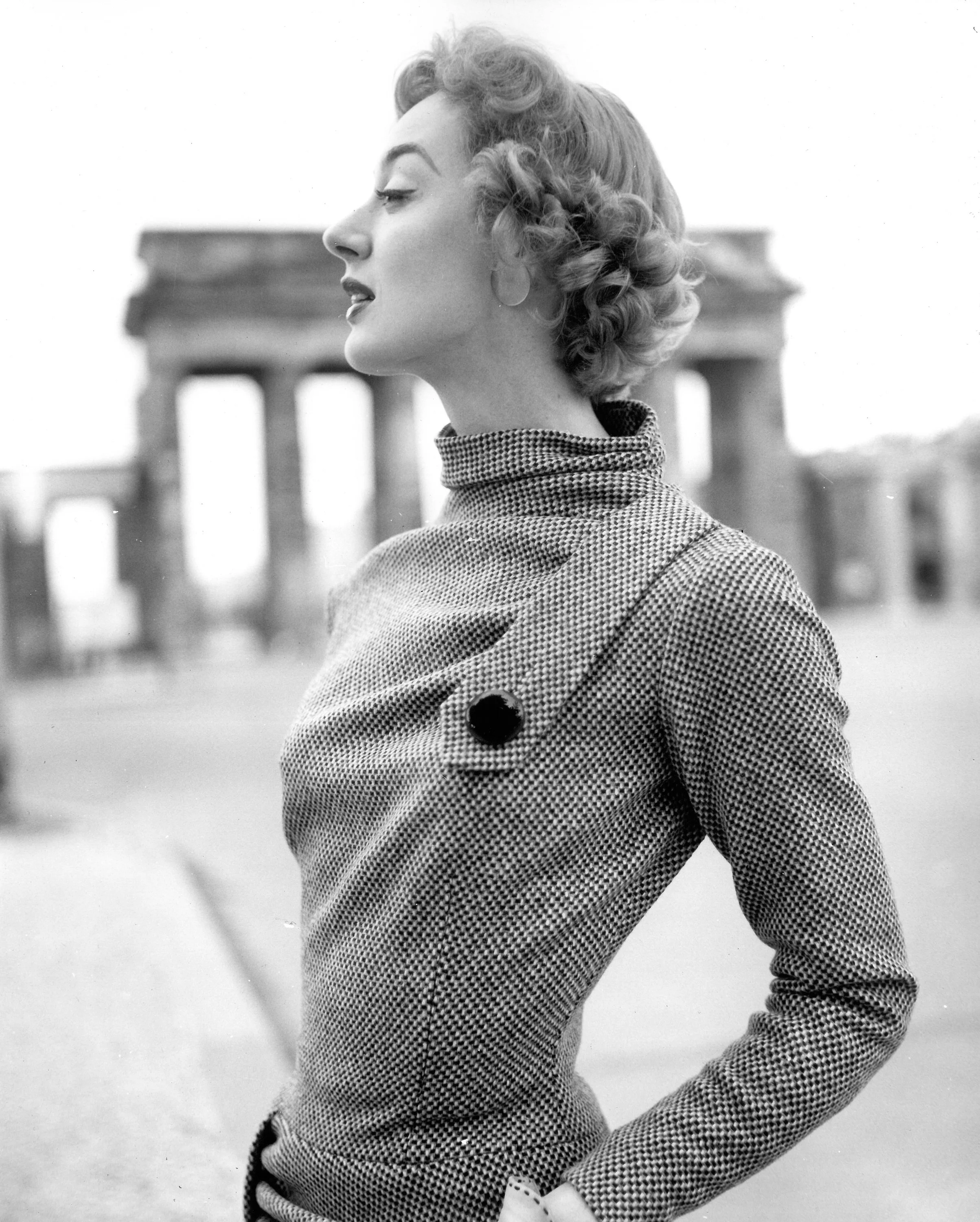 Schwarz-Weiß-Foto einer Frau im Profil vor dem Brandenburger Tor in Berlin, trägt einen hochgekrempelten Mantel und schaut nach links.