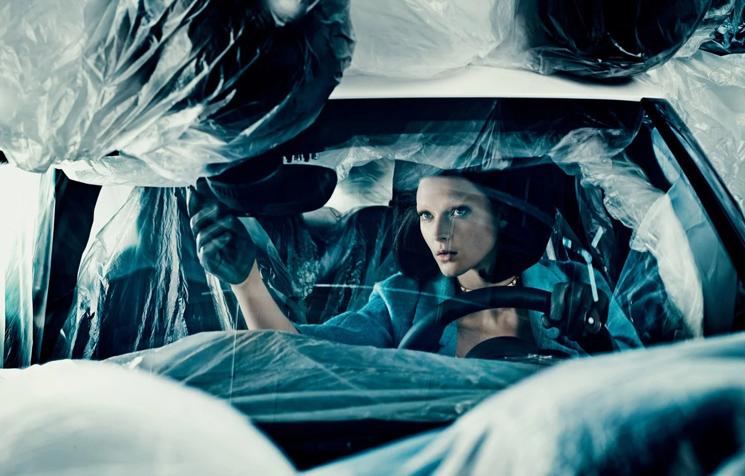 A woman with black hair looking through a windshield covered in plastic, holding a device, with a focused expression on her face.