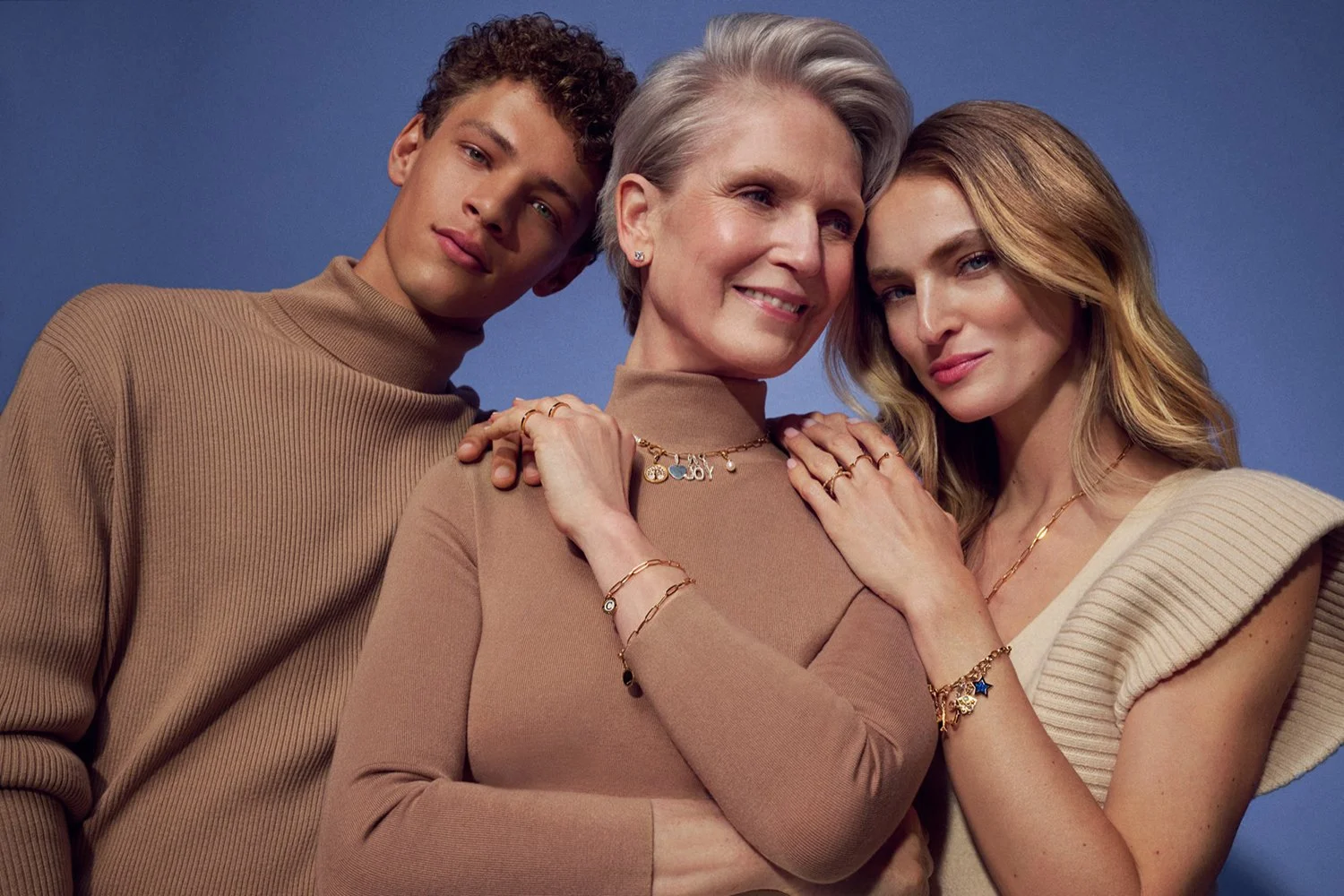 Three women, one elderly with gray hair and two younger women with blonde and brown hair, posing closely together against a plain blue background, all wearing coordinating beige and cream-colored sweaters and jewelry, smiling softly.