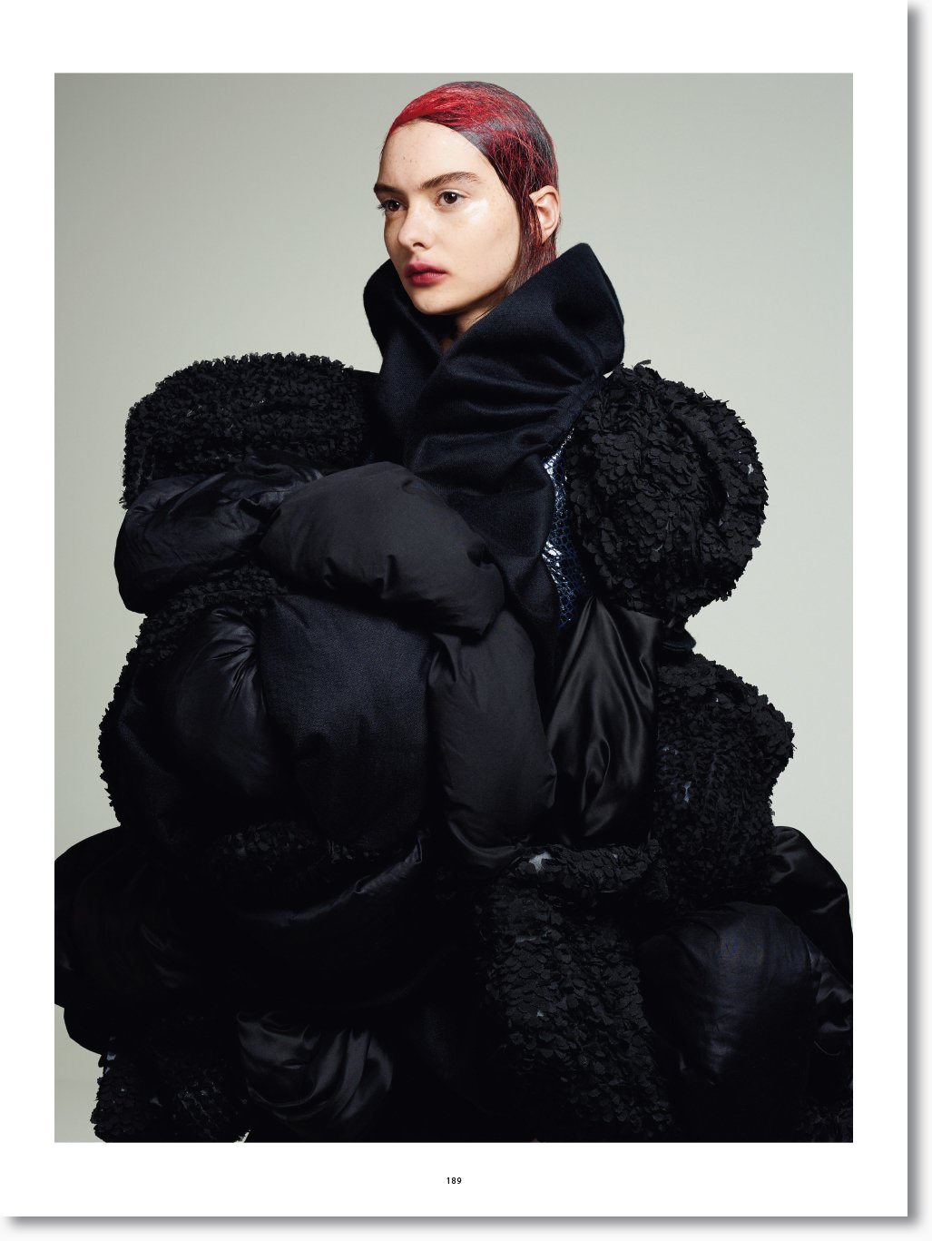 Frau trägt eine schwarze, auffällige Jacke mit großen Schulterpartien und unterschiedlichen Texturen, dunklem Make-up, rotem Haar und einem neutralen Hintergrund.