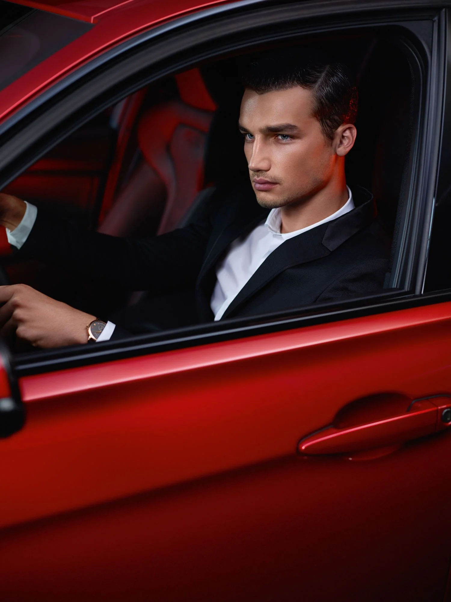 A young man with dark hair, dressed in a black suit and white shirt, sitting in a red car, looking serious.