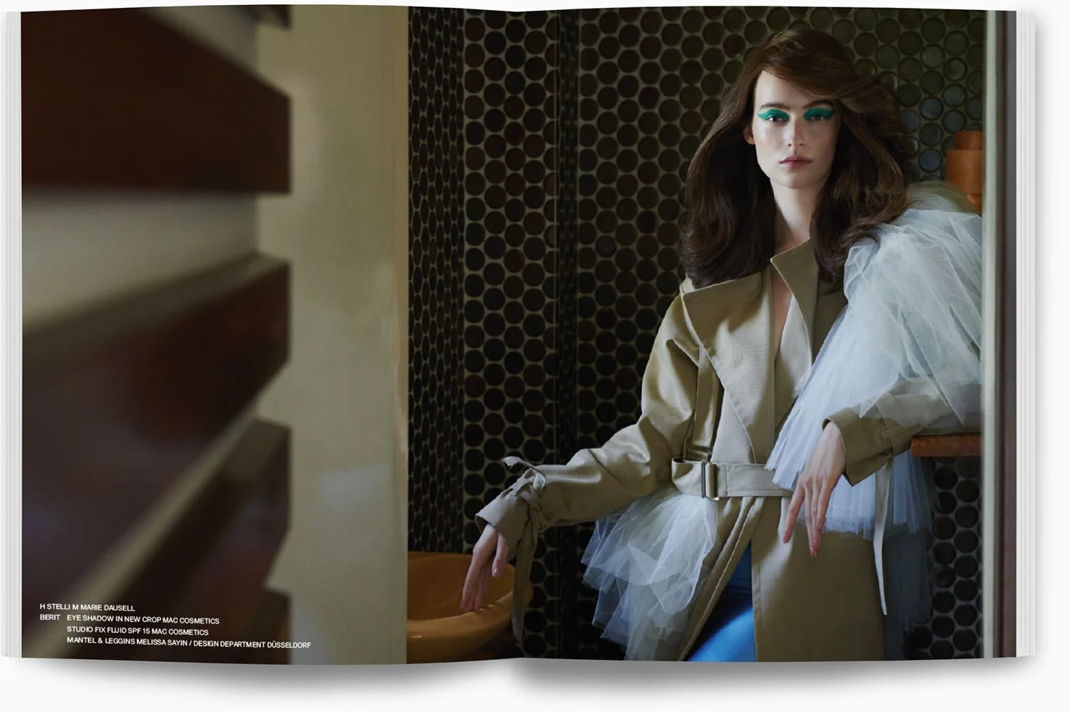 Frau steht vor einem Spiegel in einem stilvollen Raum mit dunklen, kreisförmigen Mustern, trägt einen beige Trenchcoat mit Tüll-Details und auffälligem türkisem Make-up.