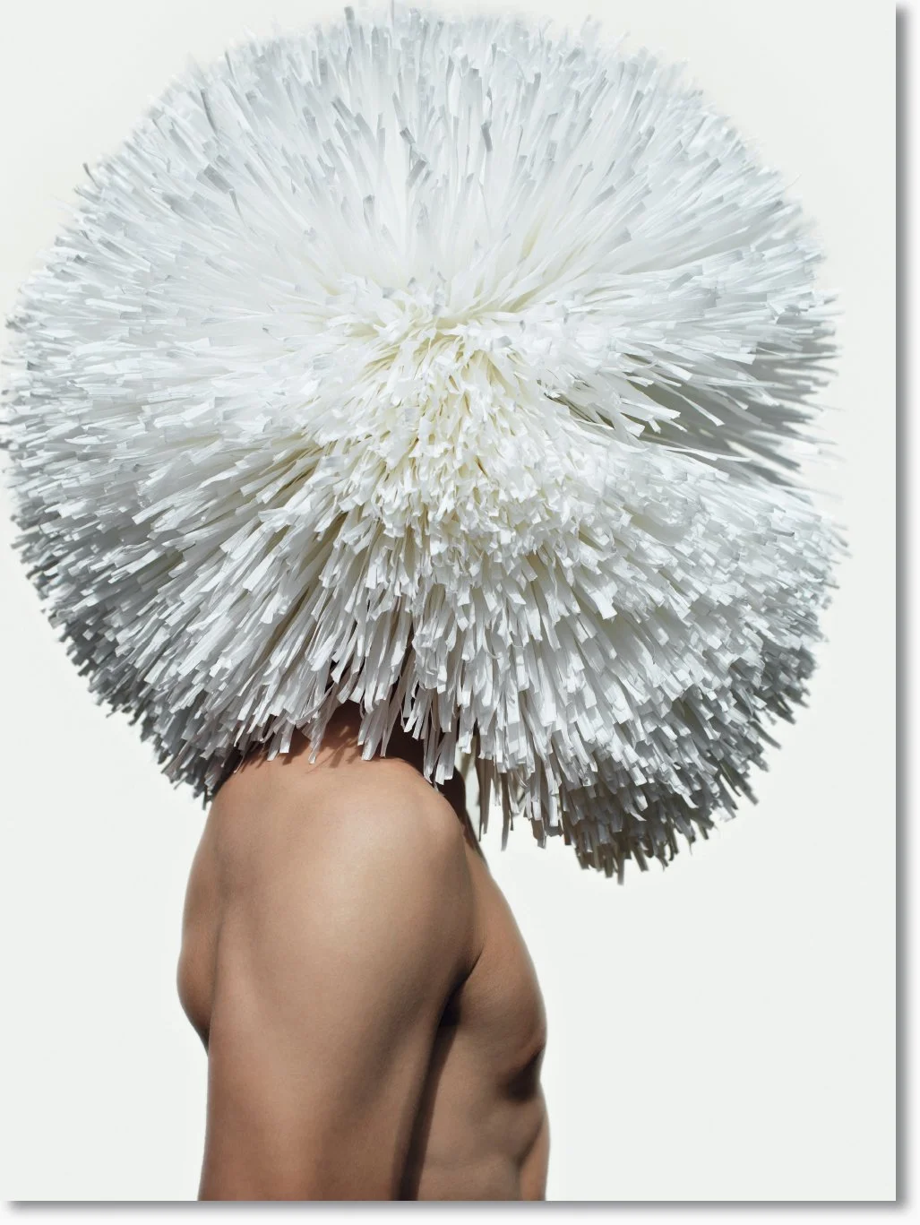 Eine flexibler, menschlicher Oberkörper trägt einen großen, weißen, fächerartigen Kunstkopf aus Papierresten.